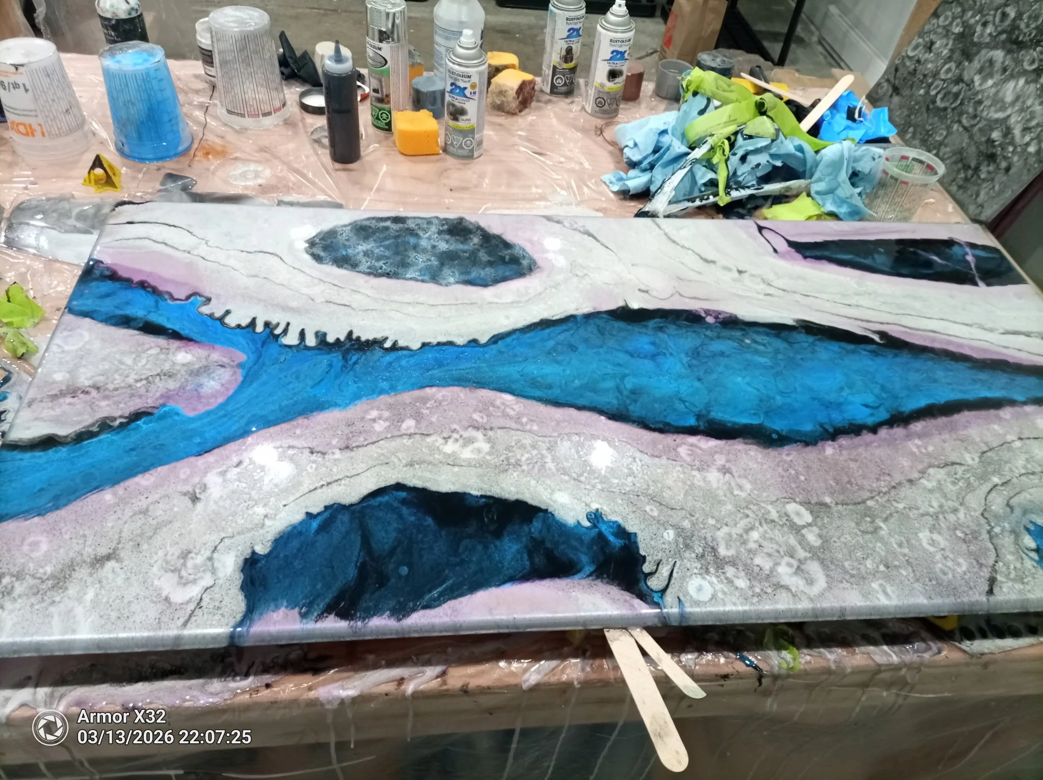 A workshop table covered with various spray paint cans, bottles of paint, and acrylic paints. The table also holds crumpled paper towels and a palette with dark and blue paint. The foreground displays a painted piece resembling a stylized skull with eyes, snout, and teeth, painted in shades of black, blue, purple, and white.