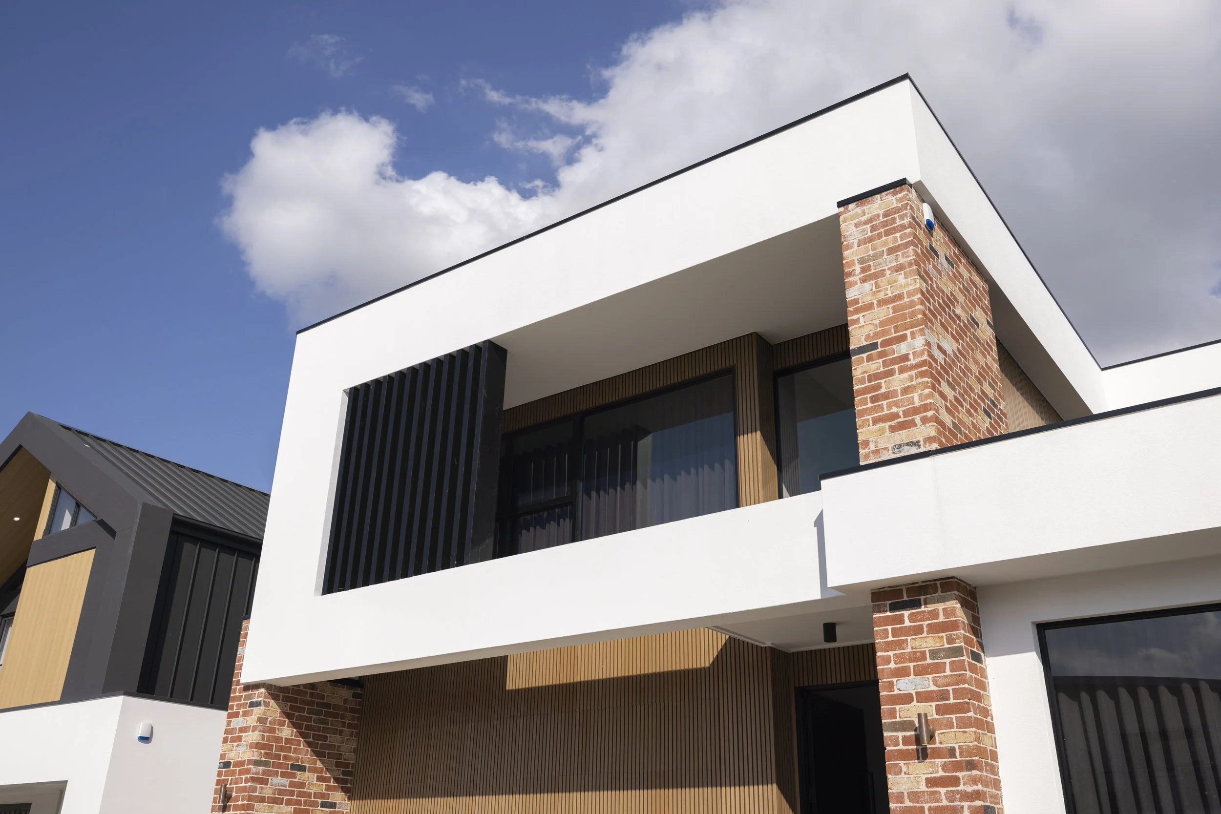 Modern multi-story house with white exterior walls, brick accents, and large windows, against a partly cloudy sky.