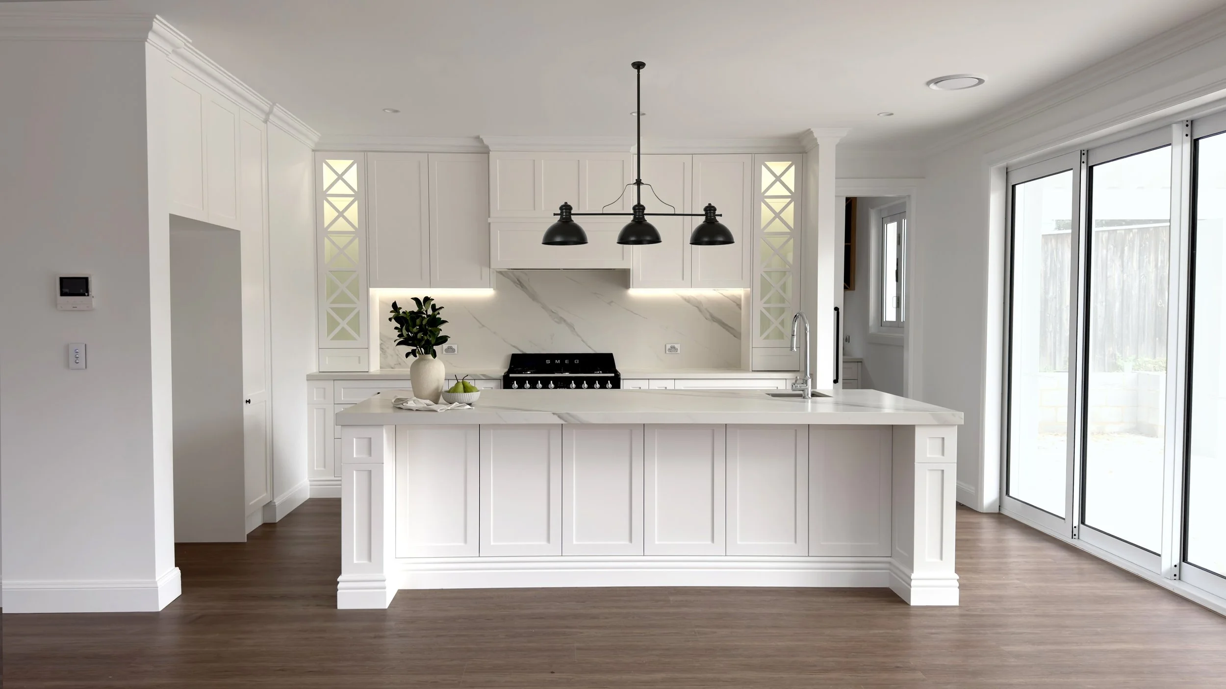 Modern white kitchen with a central island, black pendant lights, marble backsplash, and sliding glass doors leading outside.