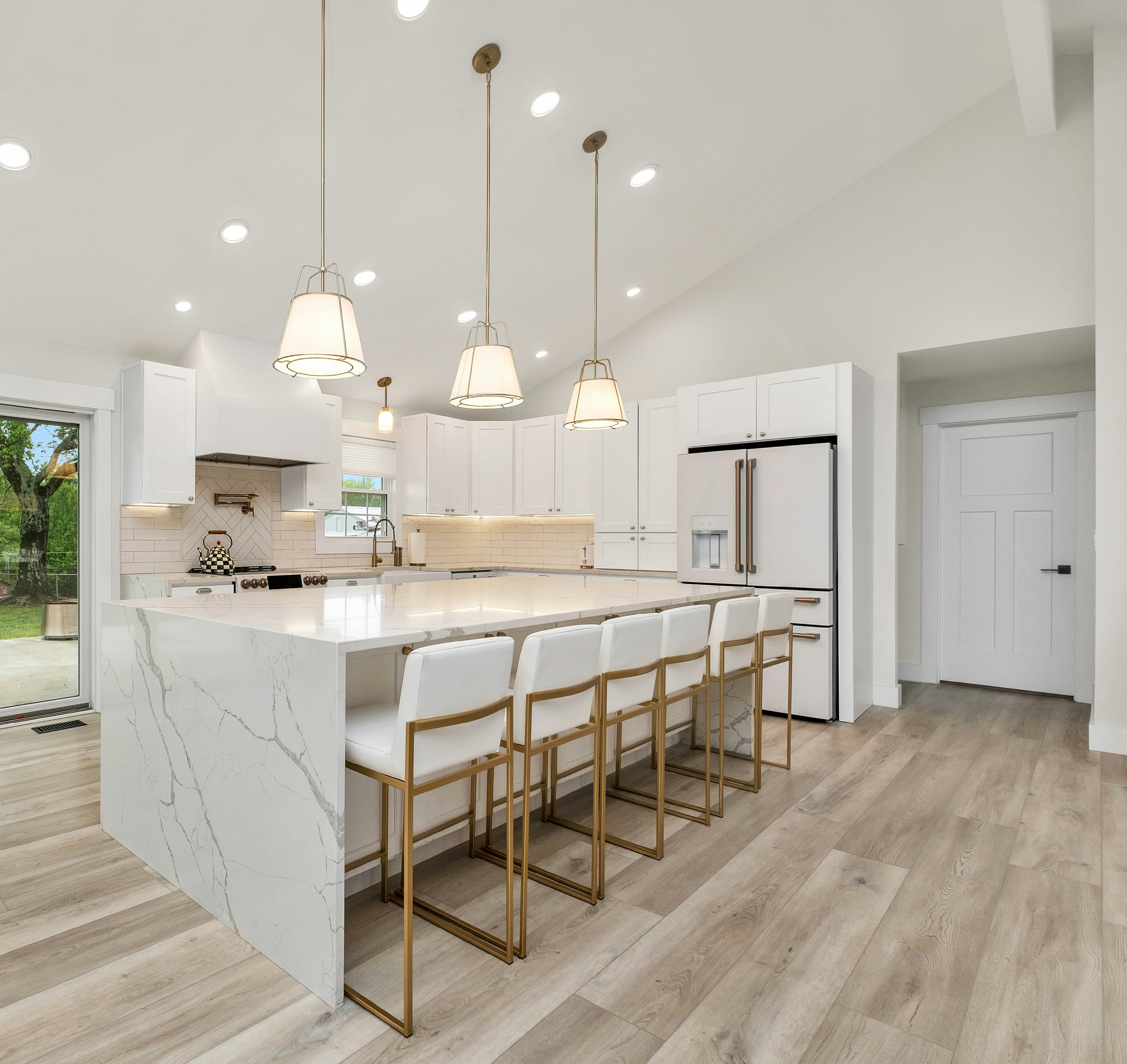 Modern white kitchen with island, black pendant lights, marble backsplash, and sliding glass doors leading outside, brown hardwood flooring, and a potted plant on the island.