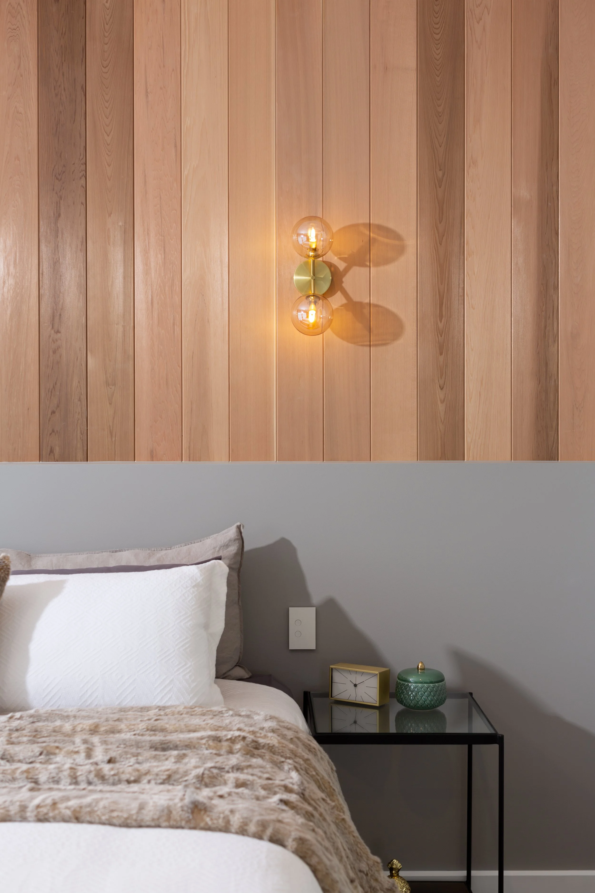 A bedroom with a wooden paneled wall, a modern wall sconce with two glowing bulbs, a white pillow, a beige textured blanket, a glass-top side table with a gold clock and a green decorative container.