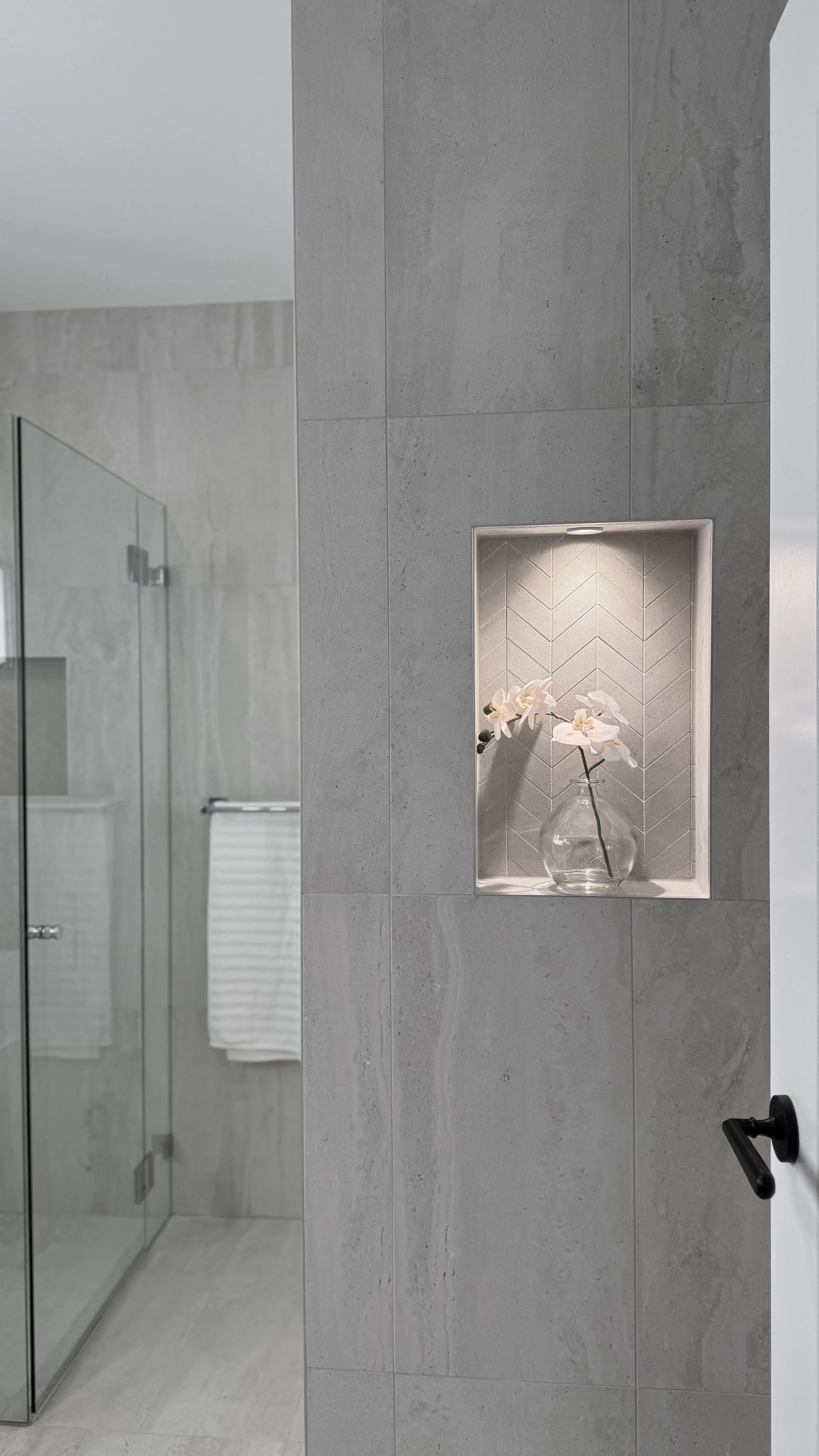 A modern bathroom with gray tiles, a glass shower enclosure, and a built-in niche with a vase of white flowers.