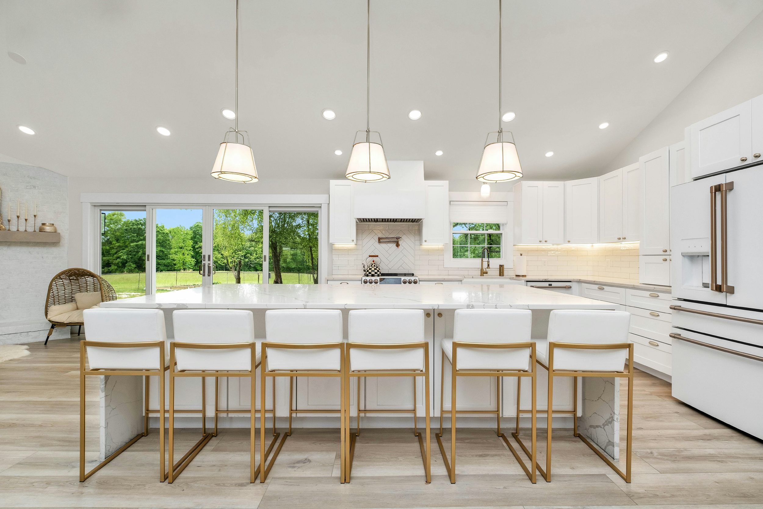 Modern white kitchen with a large island, six white and gold barstools, white cabinetry, and a view of green trees through sliding glass doors.