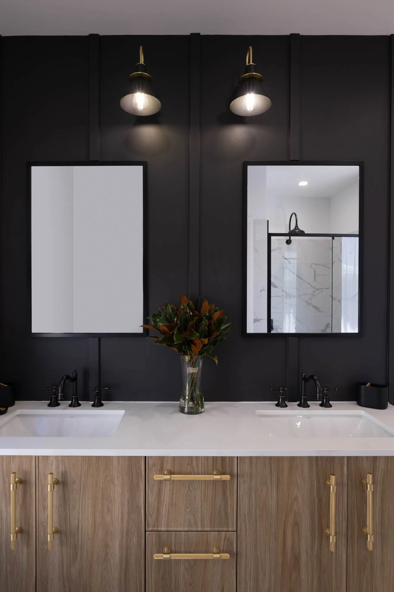 Modern bathroom with black wall, two mirrors, black fixtures, and a white countertop with a vase of green and brown leaves.