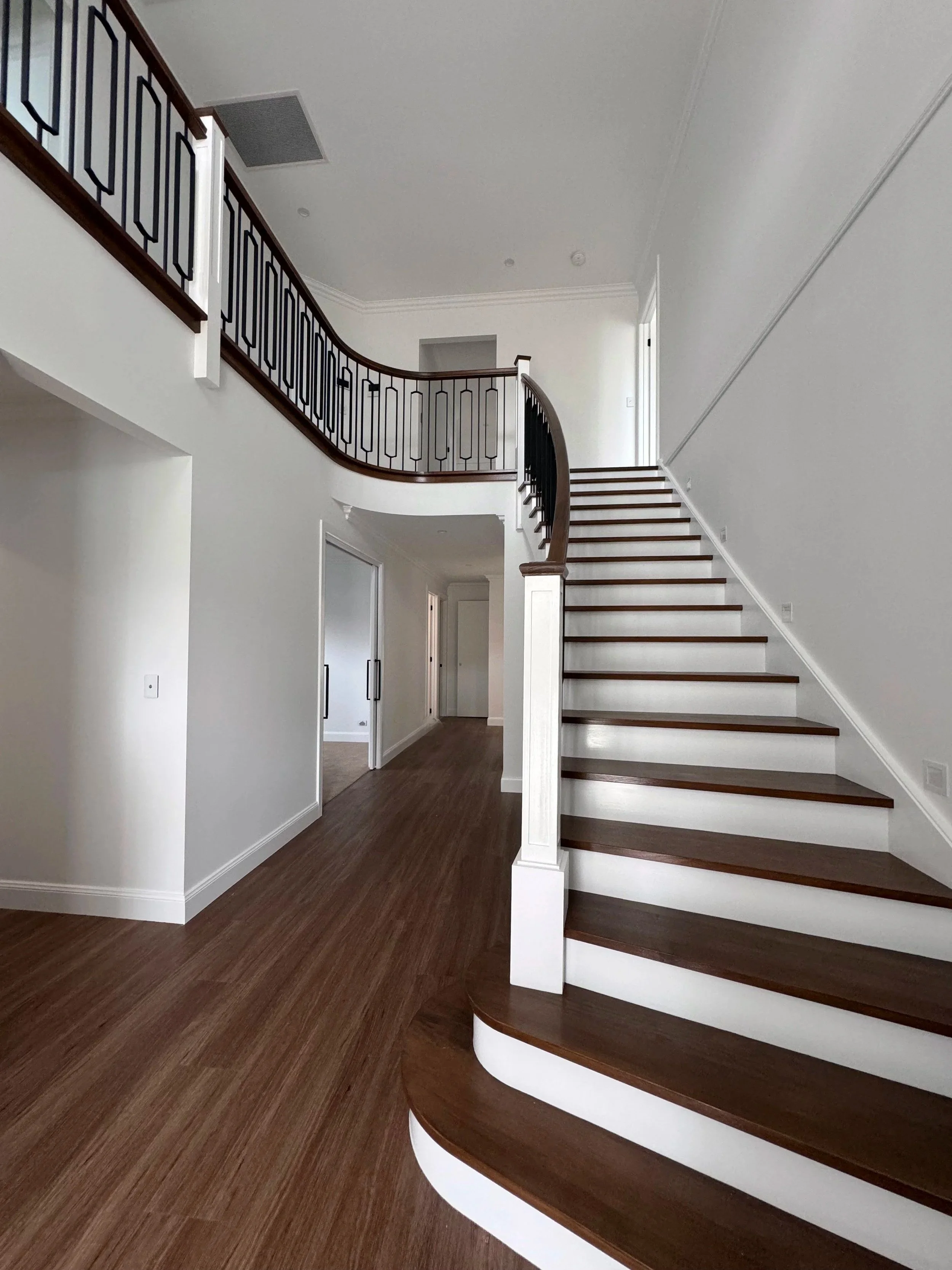 Interior view of a house with a curved staircase featuring dark wood treads and white risers, a matching dark wood and black metal railing, white walls, and wood flooring.