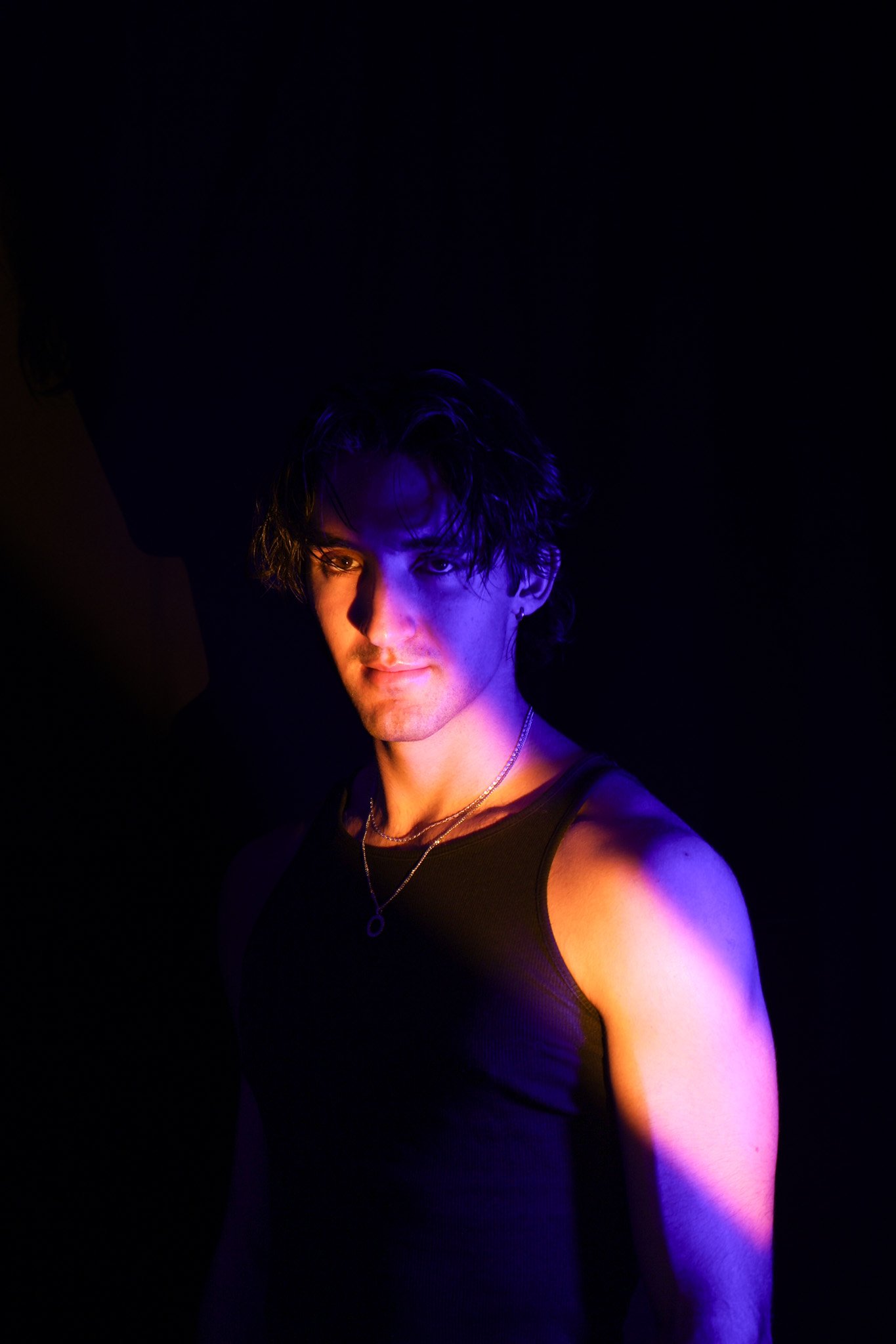A young man with dark, wavy hair and light skin is illuminated by purple and orange lighting, wearing a black tank top and a necklace, against a dark background.
