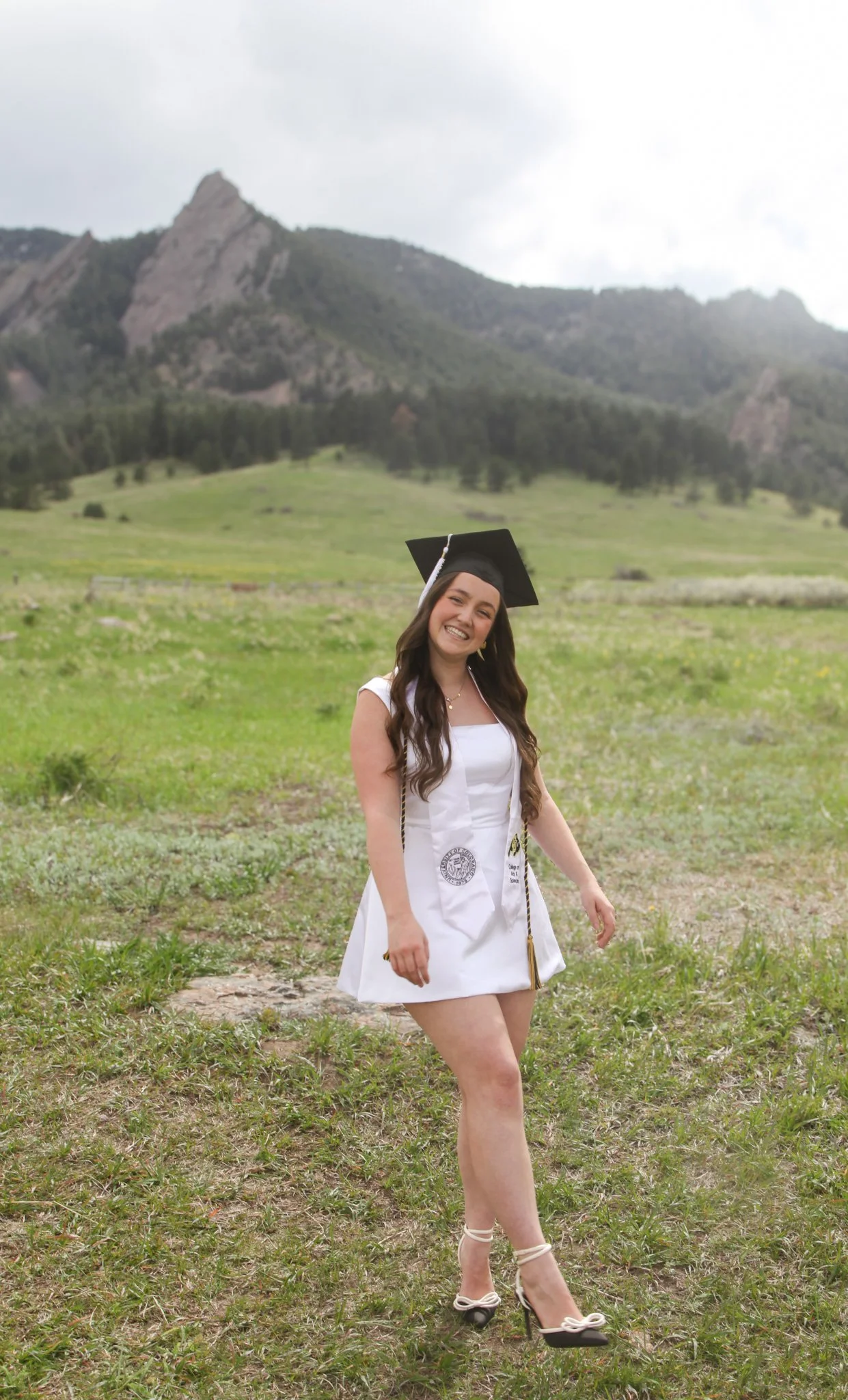 A young woman in a graduation cap and gown standing on a grassy field with mountains in the background, smiling.