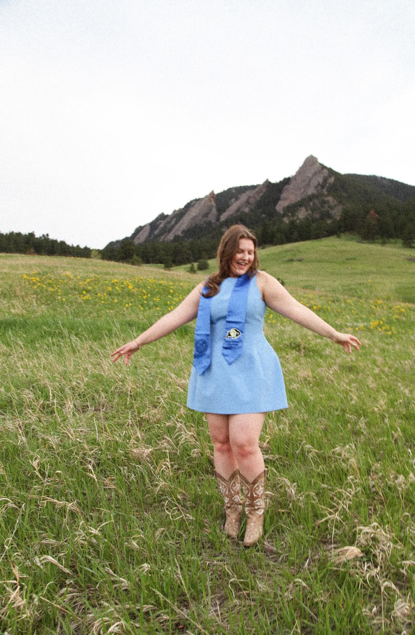 A woman in a blue dress and cowboy boots standing in a grassy field with mountains in the background, smiling with arms outstretched.
