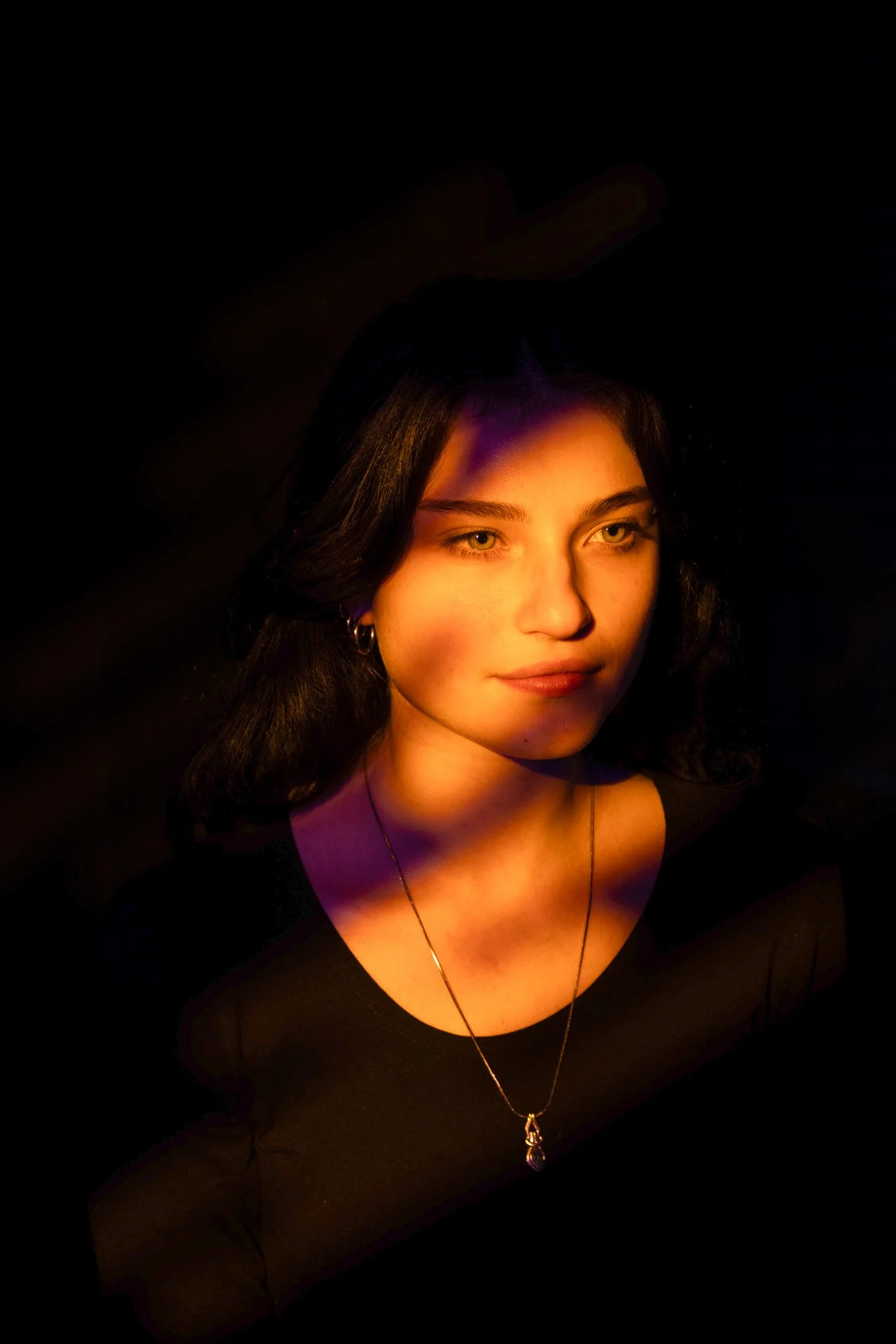 A young woman with dark hair, wearing a black top and a necklace, is illuminated by a warm, orange light against a dark background.