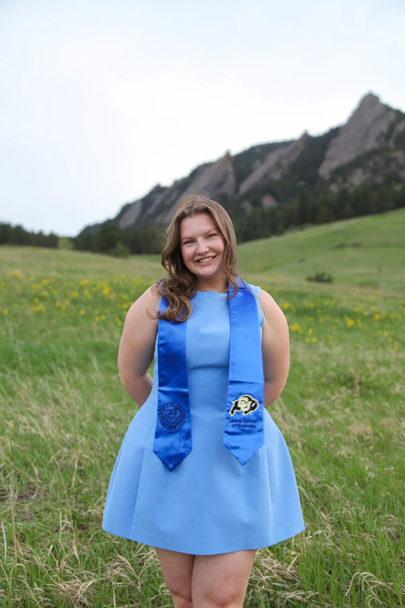 A young woman in a blue dress and a blue graduation stole standing outdoors in a grassy field with mountains in the background, smiling.