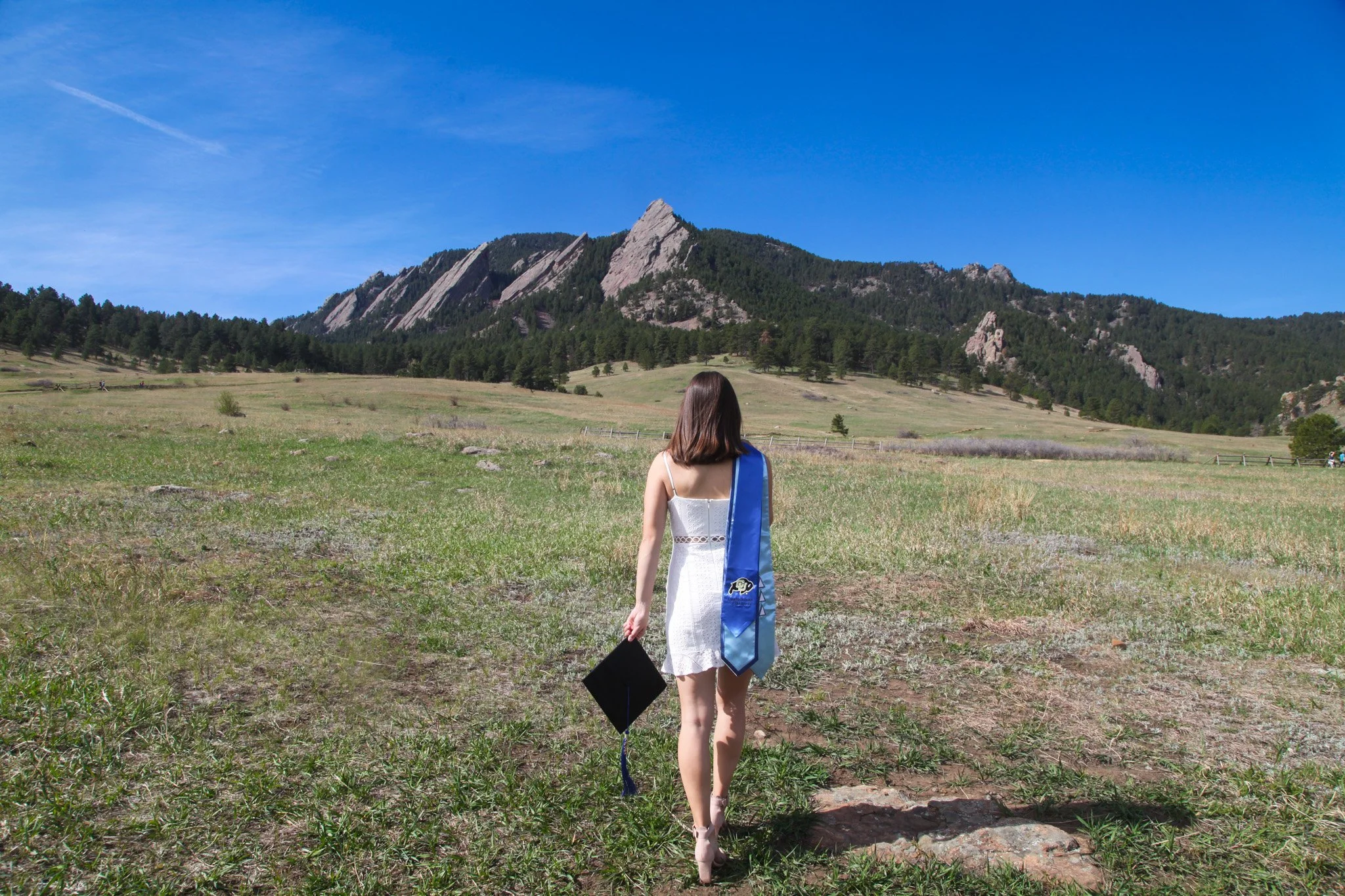 A woman in a white dress holding a graduation cap, walking towards a mountain landscape under a clear blue sky.