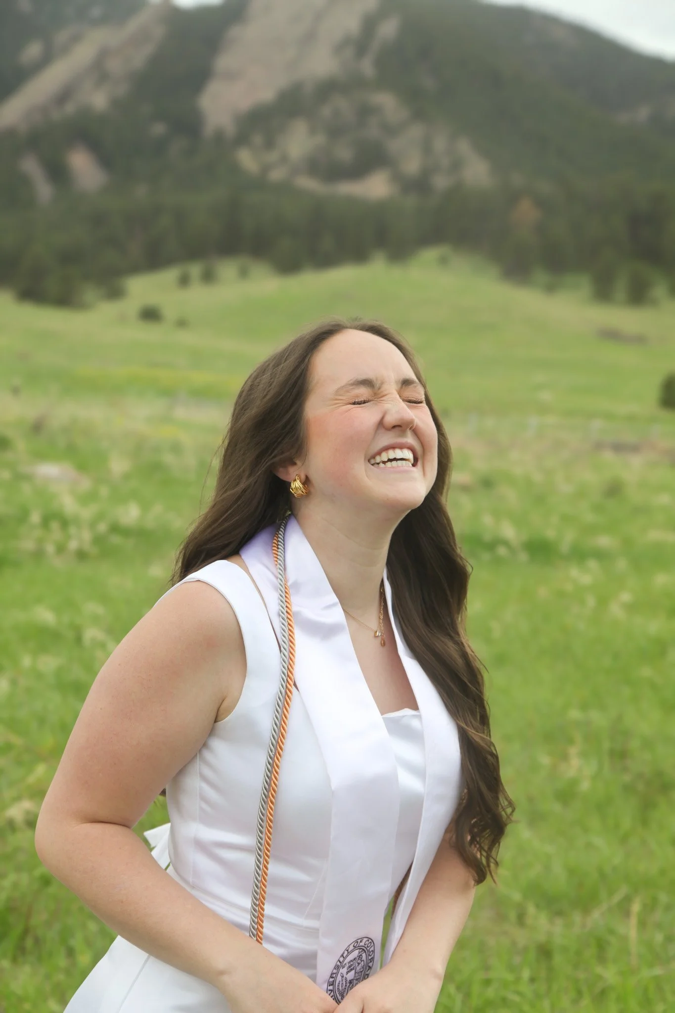 A young woman in a white graduation dress, smiling and closing her eyes, standing outdoors with green grass and mountains in the background.