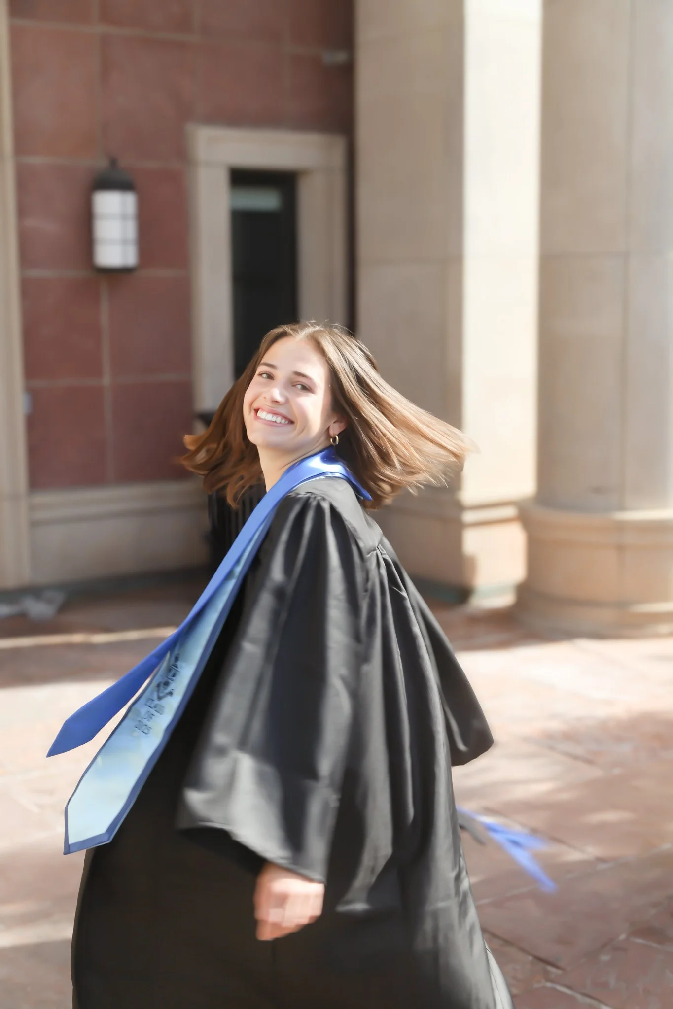 A young woman in a graduation gown and blue stole smiling and turning her head outdoors in front of a building with columns.