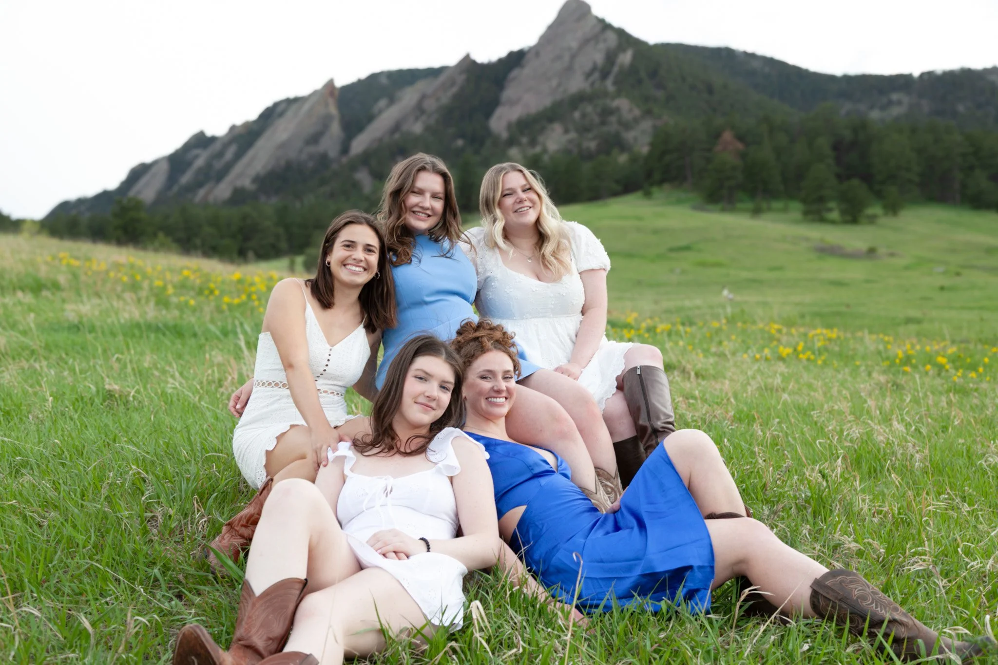 Six young women sitting and lying on a grassy field with mountains in the background, dressed in summery outfits and smiling at the camera.