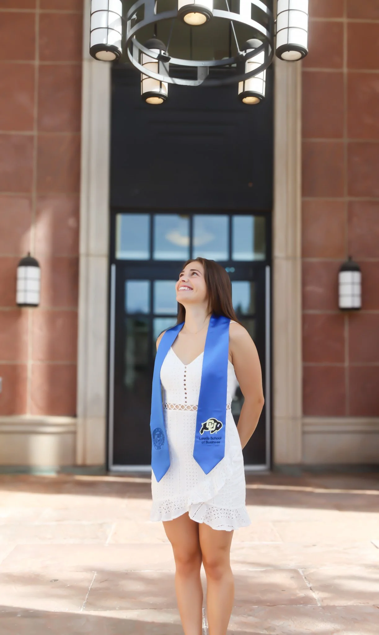 A young woman in a white dress with a blue graduation stole standing outside in front of a building, smiling and looking upward.