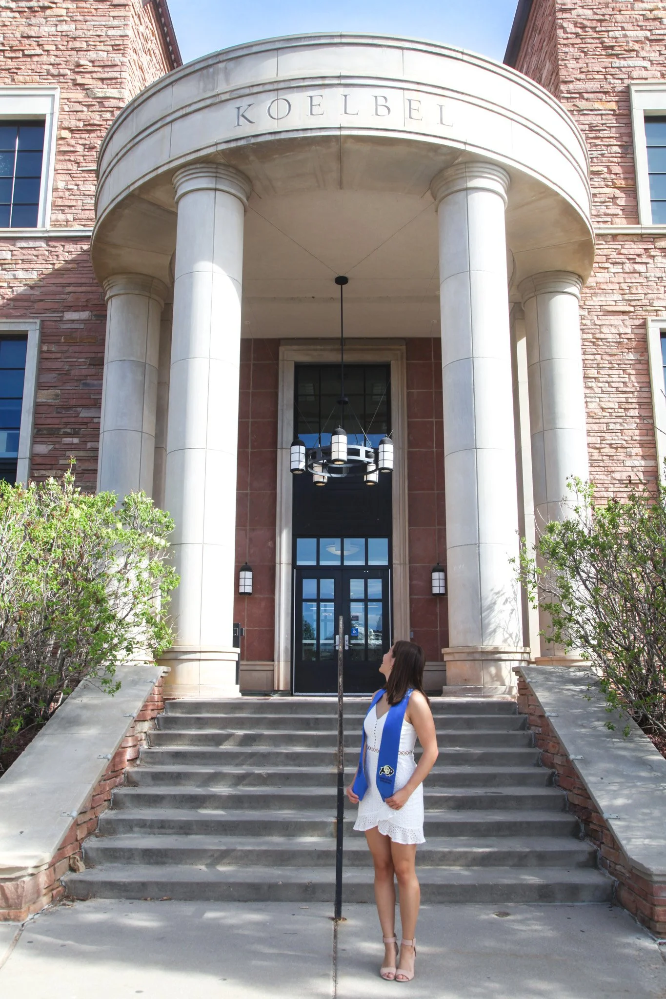 A woman in a white dress and heels wearing a blue graduation stole stands on the steps of a building with the word 'KOELBEL' on the curved entrance. The building has brick walls, large pillars, and modern lighting fixtures.