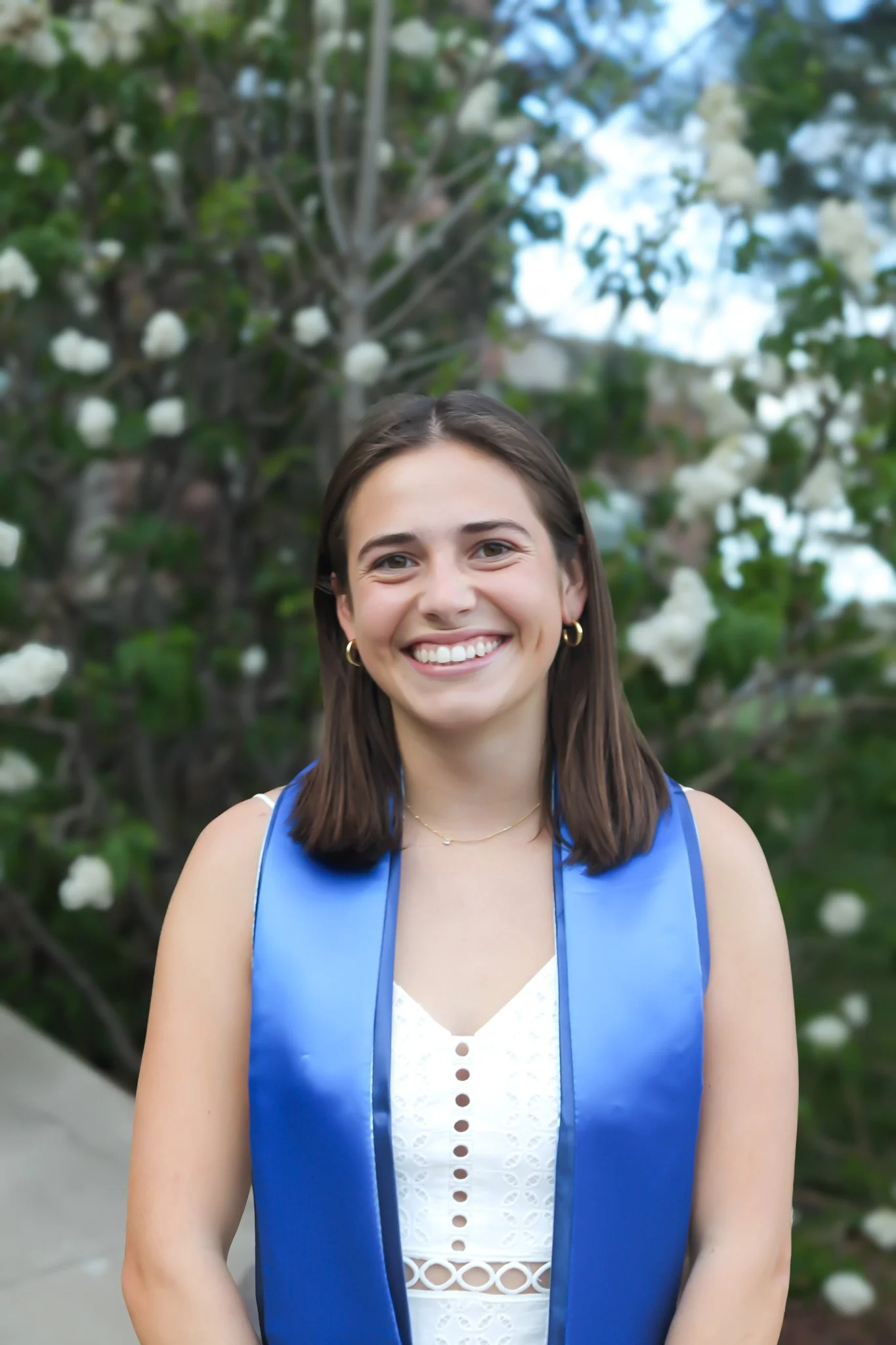 A young woman with shoulder-length brown hair, smiling, wearing a white dress and blue graduation stole, standing outdoors in front of a bush with white flowers.