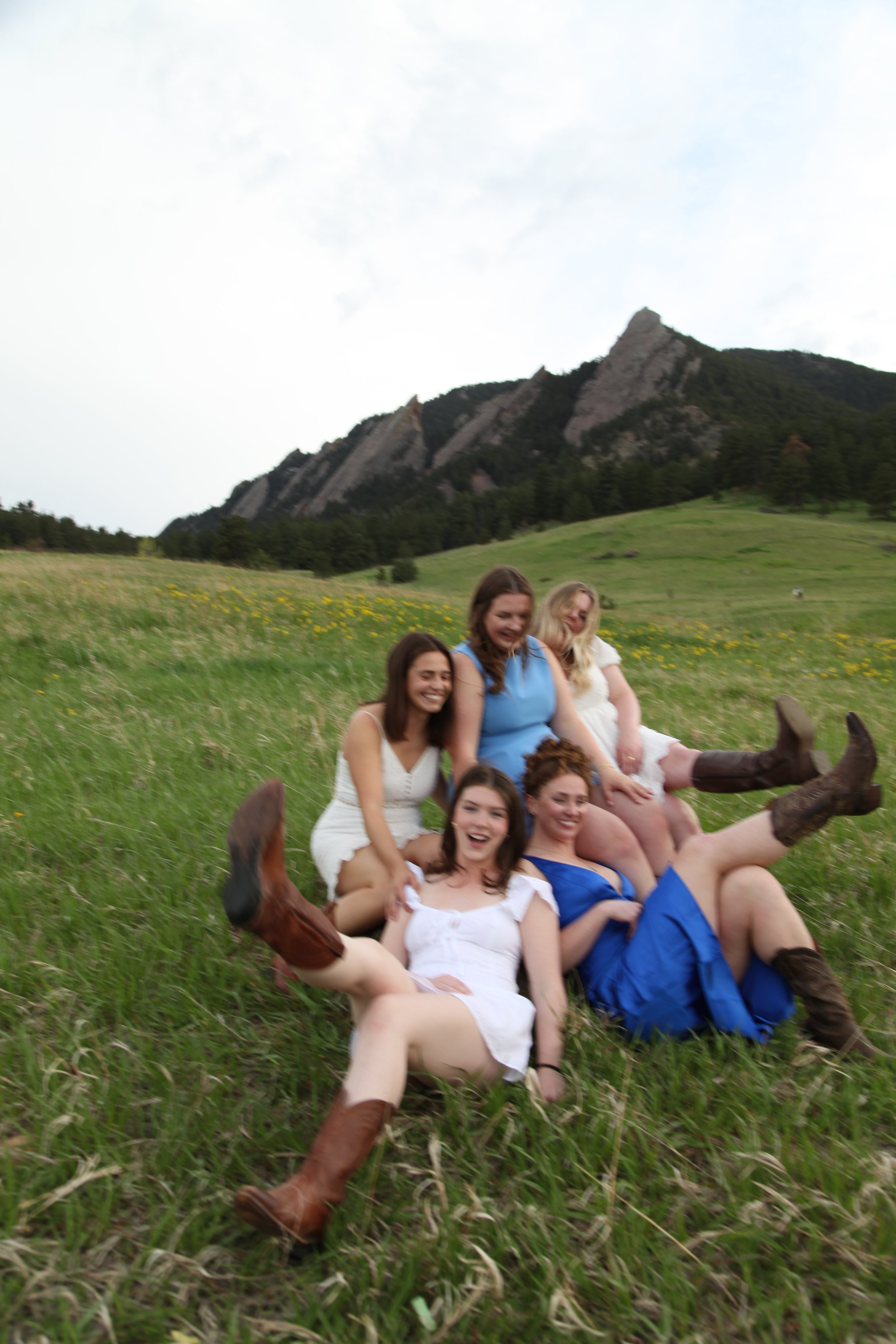 Group of six women in colorful dresses and cowboy boots sitting and lying on a grassy field with a mountain in the background.