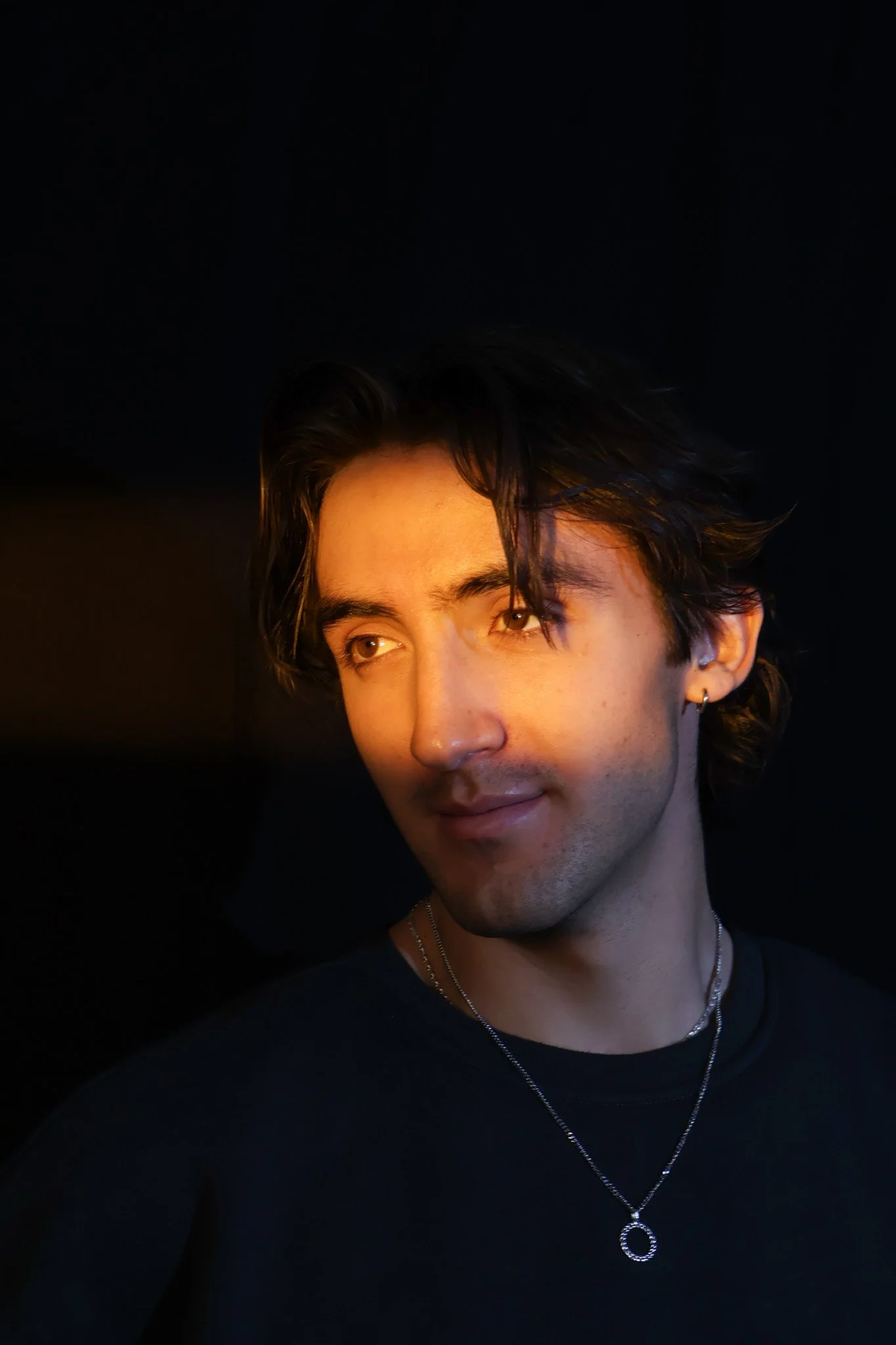 Portrait of a young man with dark hair, earrings, and necklaces, looking to the side with a warm light on his face against a dark background.