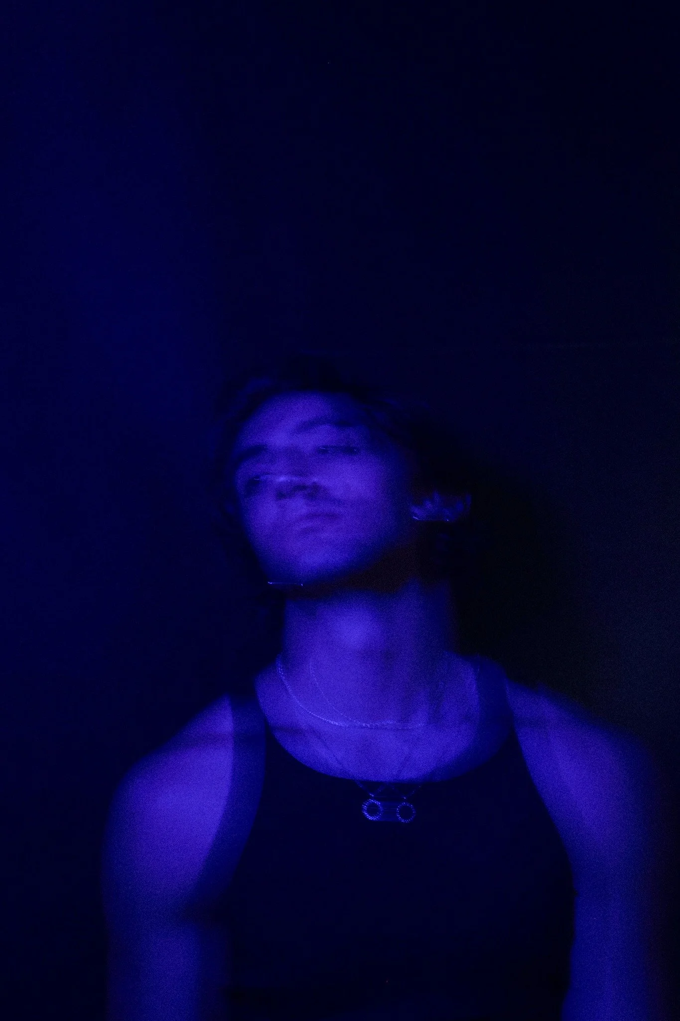 Person with short hair and earrings in a dark, blue-lit environment, wearing a black top and layered necklaces.