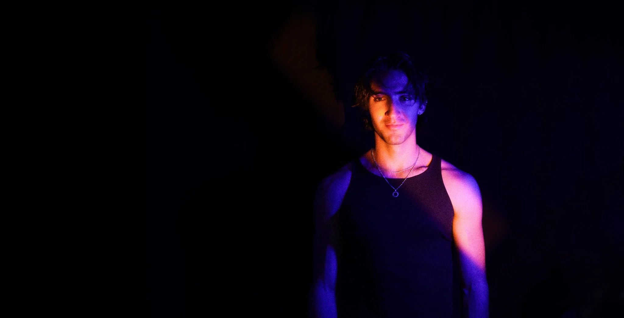 Young man with curly hair wearing a black tank top, illuminated by purple and blue lighting against a dark background.