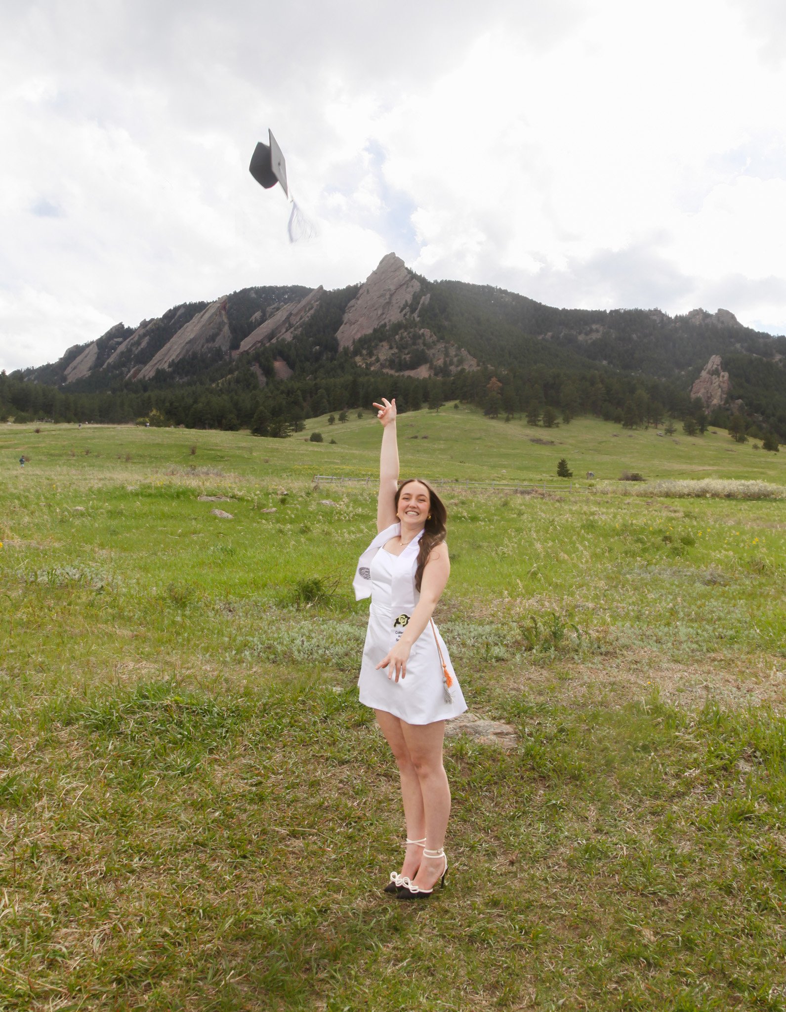 A young woman in a white dress and high heels celebrating her graduation outdoors in a grassy field with mountains and trees in the background, tossing her black graduation cap in the air and smiling.