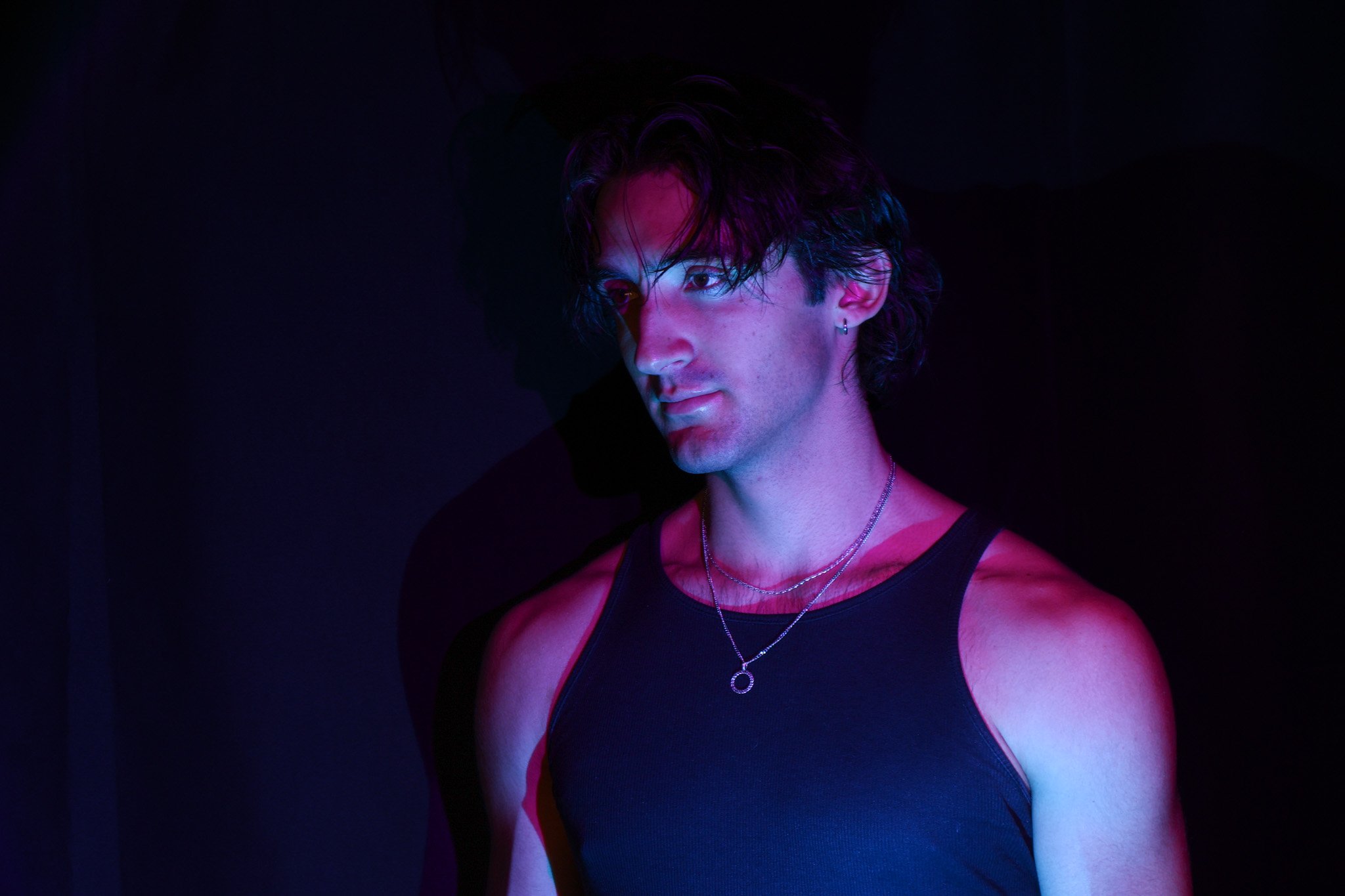 A young man with curly hair and earrings wearing a black tank top, illuminated by pink and blue lighting, against a dark background.
