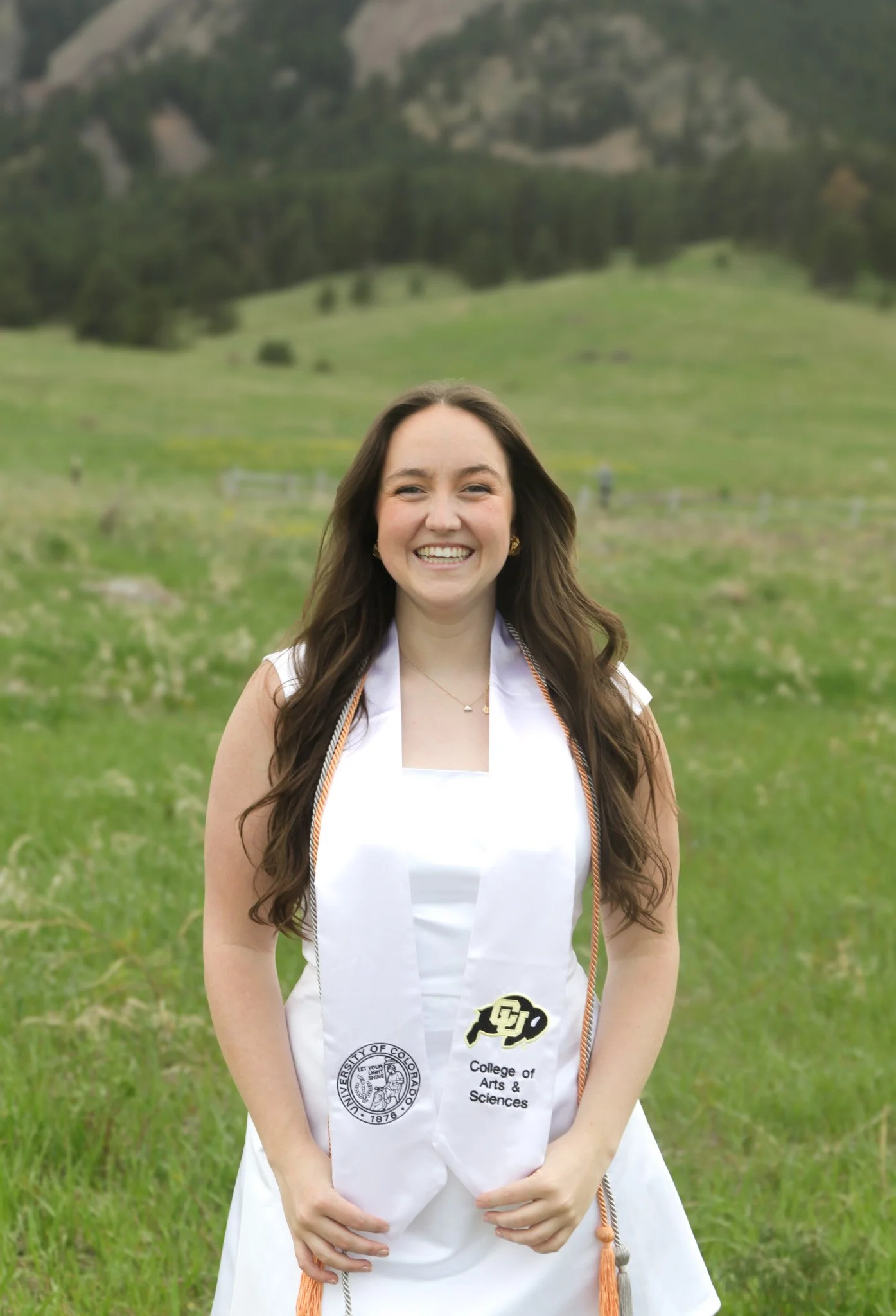 A young woman in graduation attire standing outdoors in a grassy field with mountains in the background, smiling.