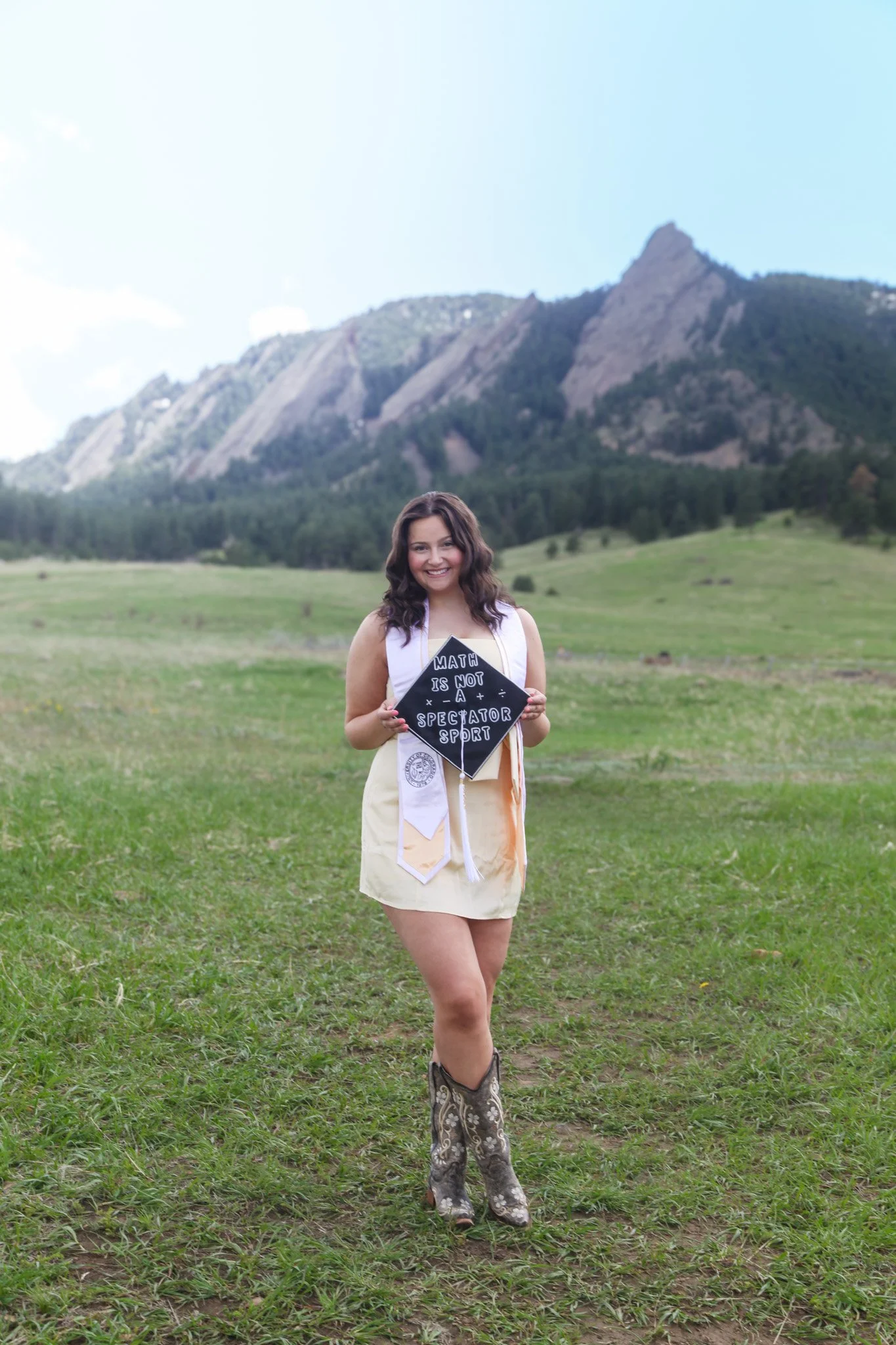 A woman standing in a green field with mountains in the background, holding a sign that reads "Math is not a spectator sport." She is wearing a yellow skirt, cowboy boots, and a white vest with medals hanging from it.