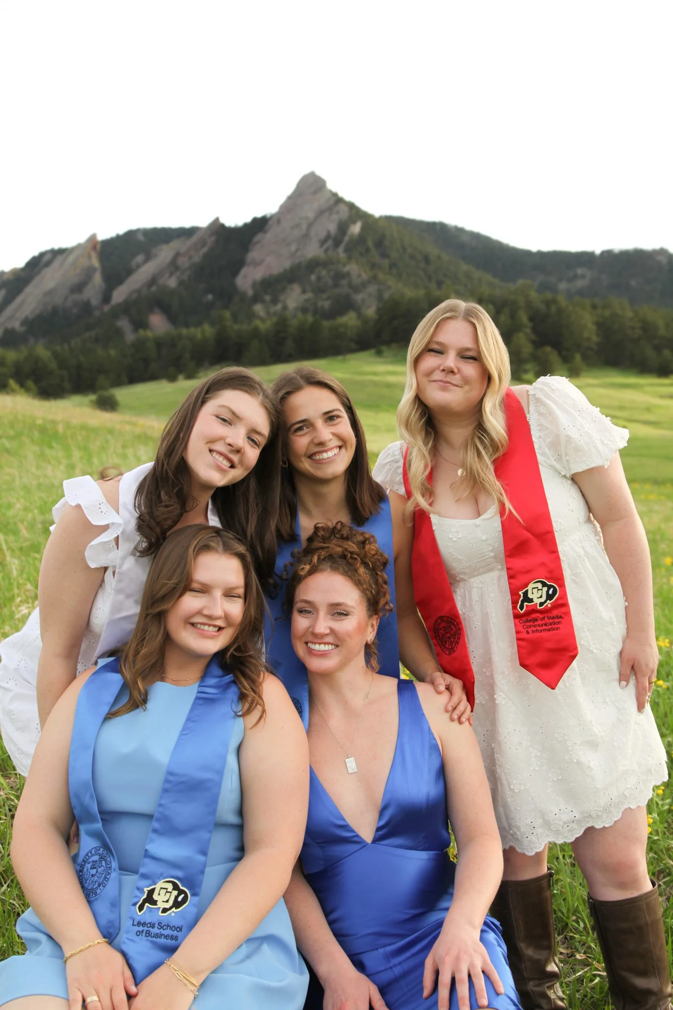 Group of five women, some in blue graduation sashes, posing outdoors in a grassy field with mountains in the background.