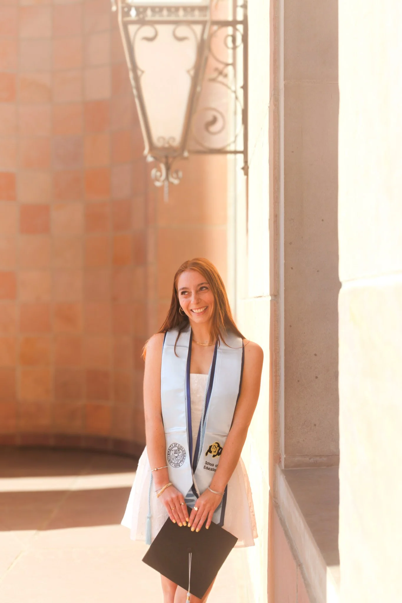 A young woman in a white dress and a graduation stole smiles while holding a diploma, standing beside a stone wall with a decorative street lamp overhead.