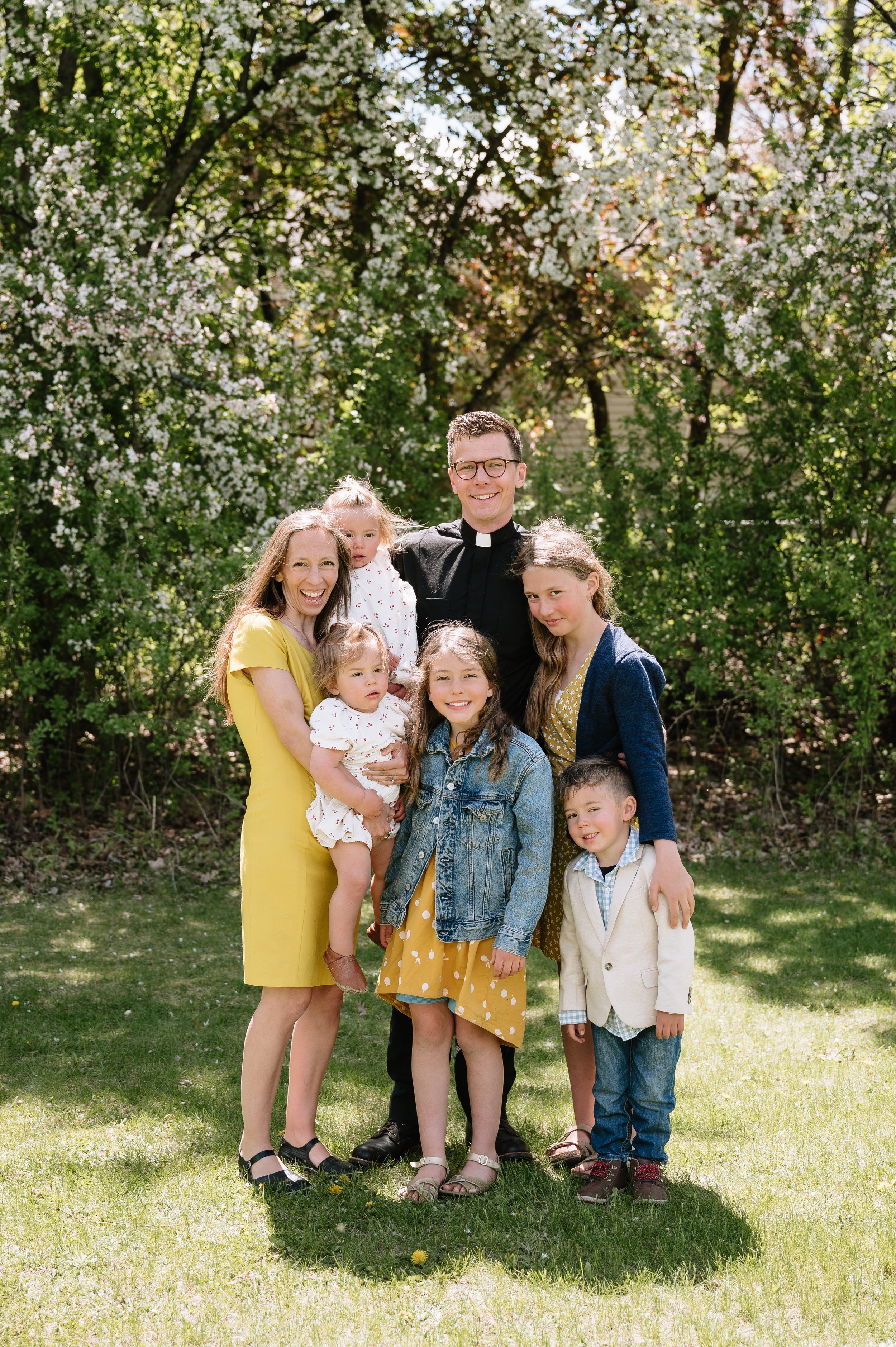 A group of seven people, including a priest and children, standing outdoors in front of blooming trees on a sunny day, smiling.