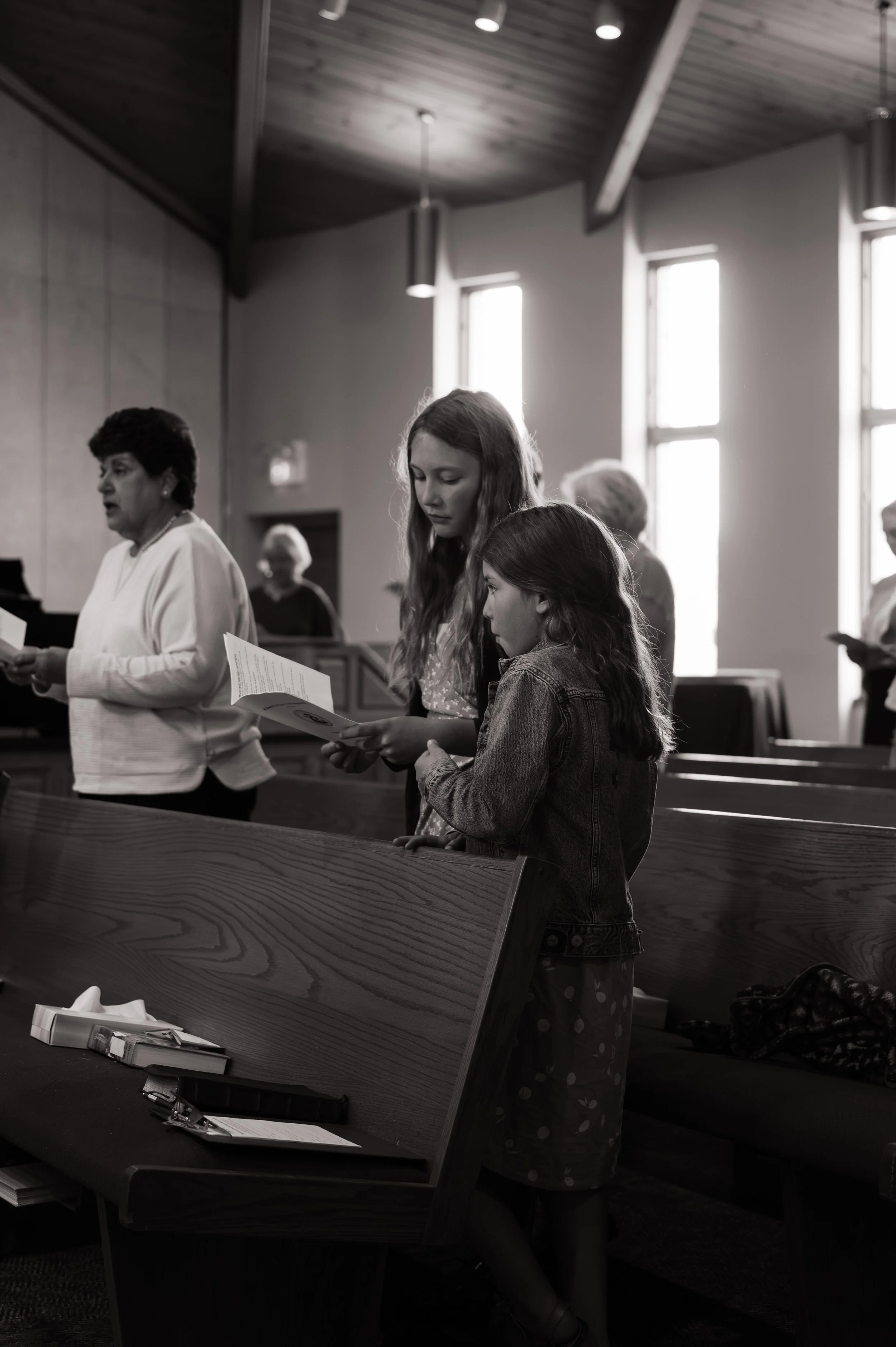 A woman and a girl are standing inside a church, holding hymnals, and singing or praying. Other people are in the background.