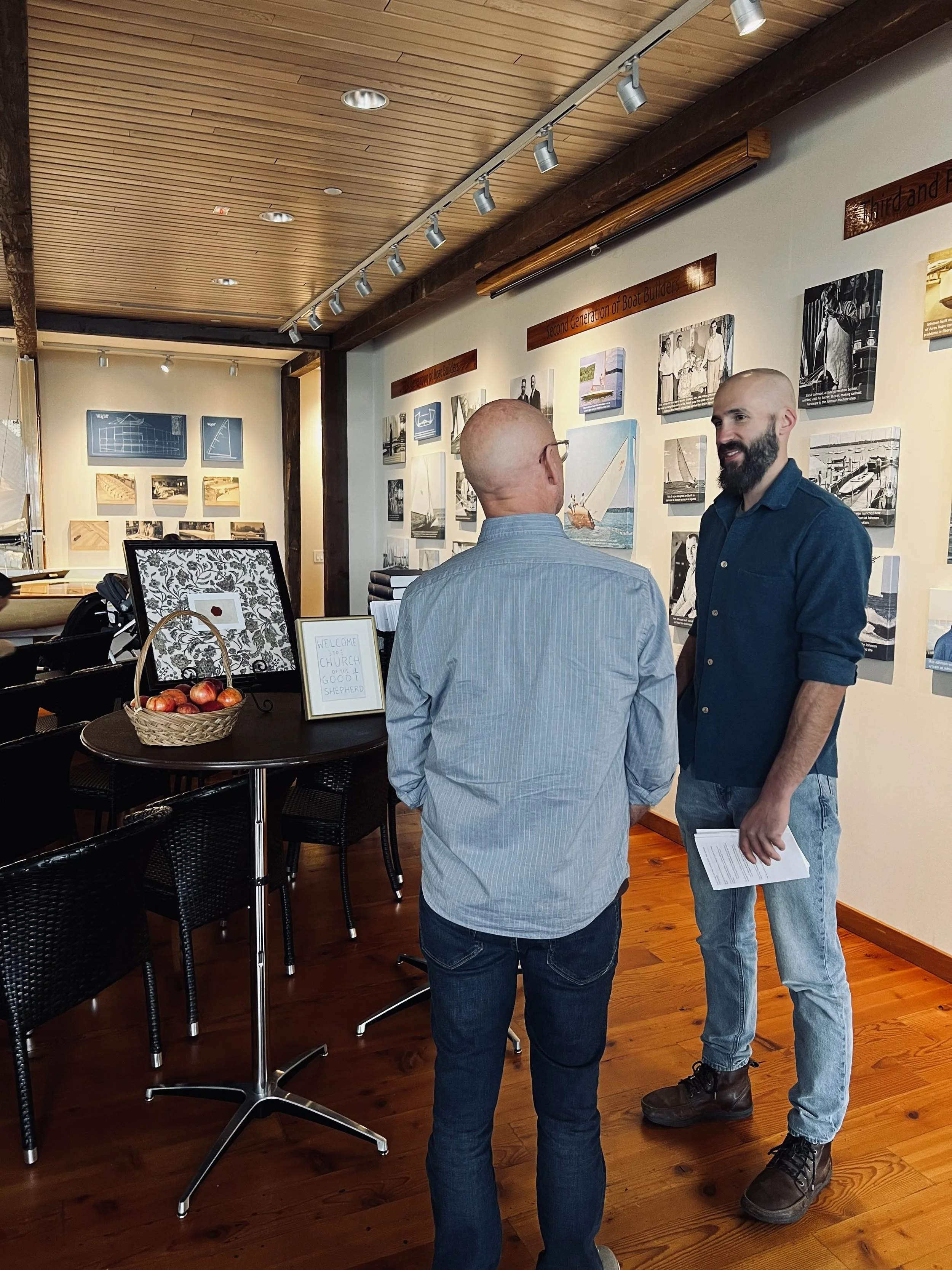 Two men are talking inside an art gallery or museum with photos and paintings on the wall. One man is bald and wearing glasses, a light blue shirt, and jeans. The other man has a beard, is bald, and wearing a dark blue shirt with rolled-up sleeves, jeans, and boots. A table with a basket of apples and framed signs is nearby.