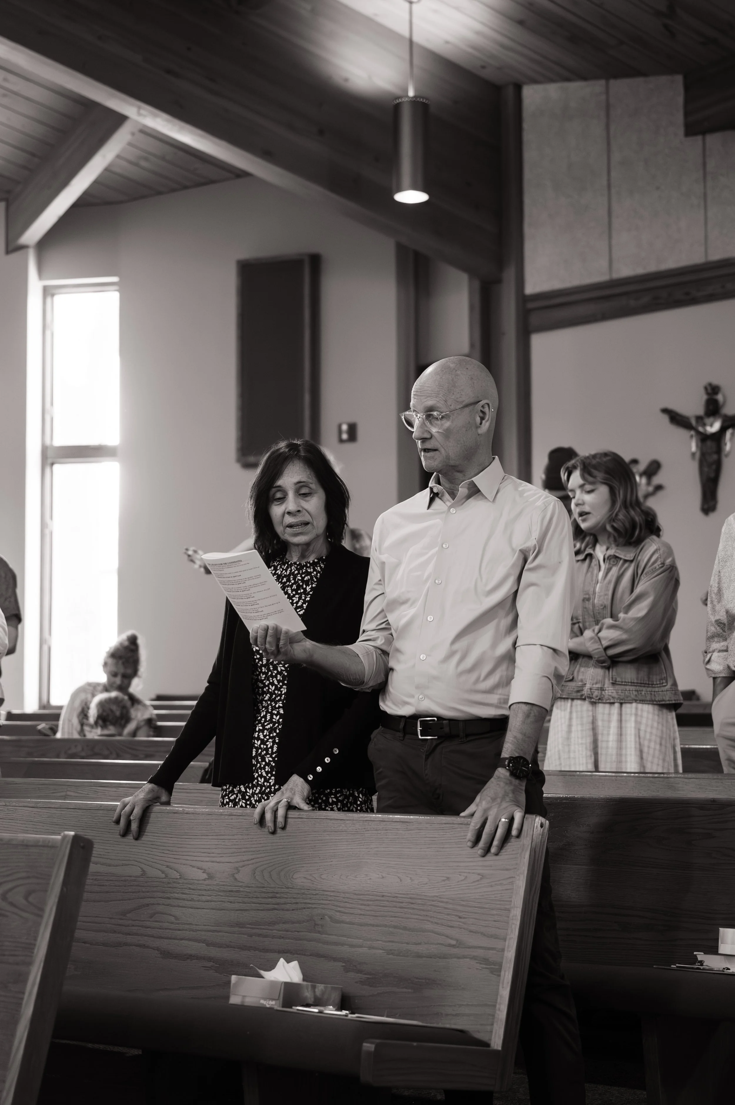 Inside a church, a man in glasses and a white shirt stands with a woman singing hymns, both with their eyes closed. Other people are visible in the background singing, with religious symbols on the wall.