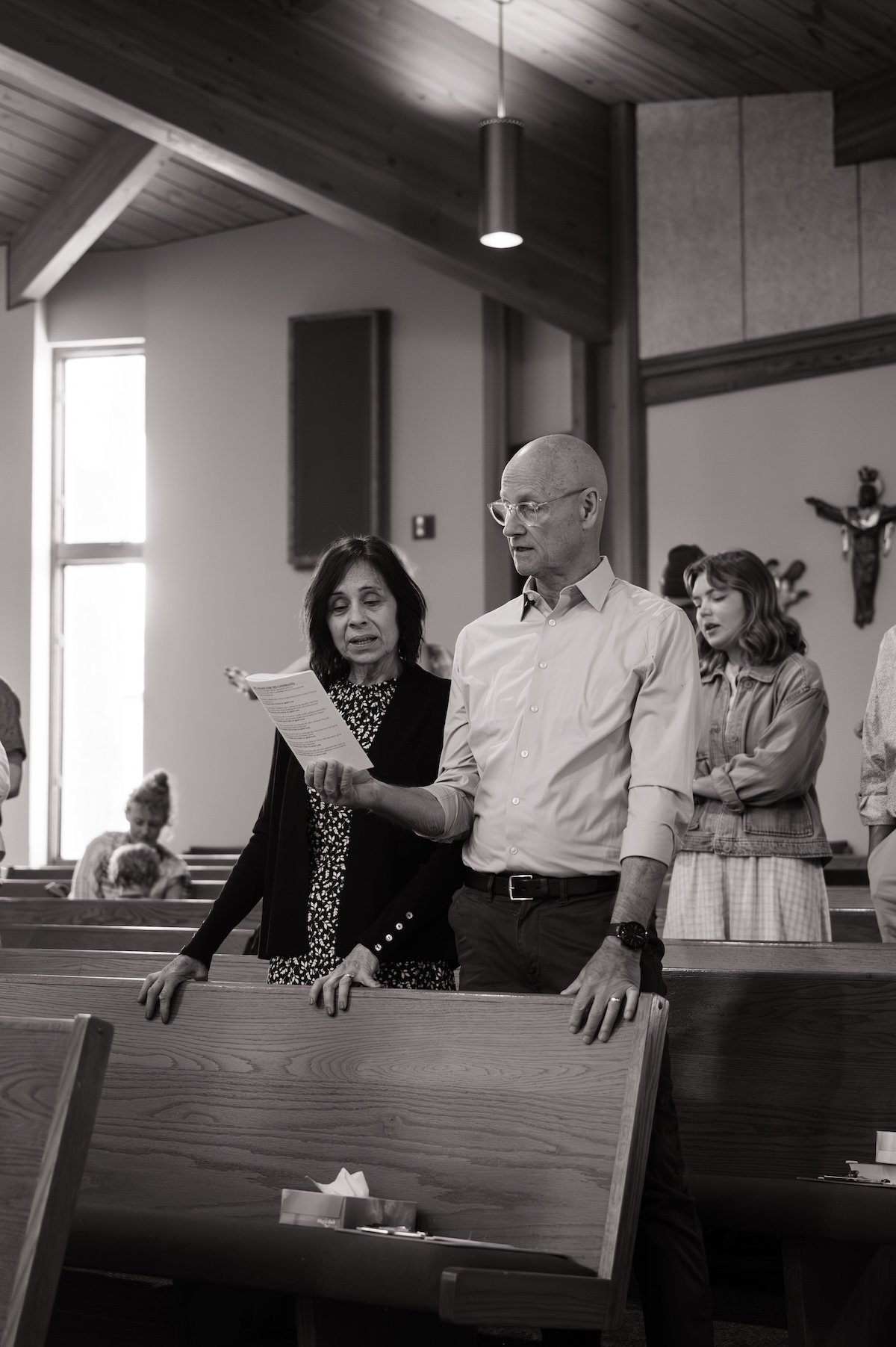 A group of people standing in a church, with a woman reading from a paper and a man praying, surrounded by others, including a young girl, during a religious service.