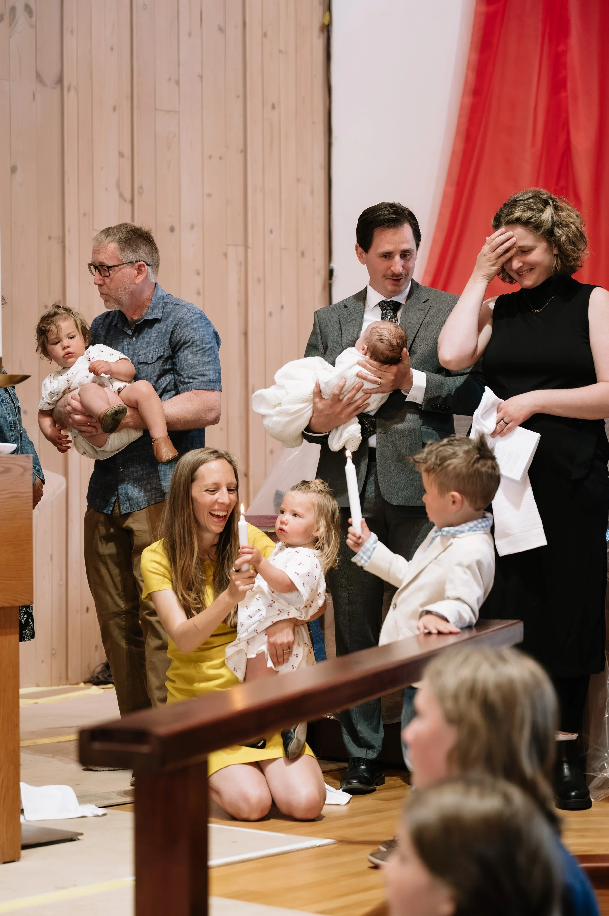 Family gathered for a religious ceremony, with children, a baby, and adults inside a church with wood-paneled walls and red curtains.