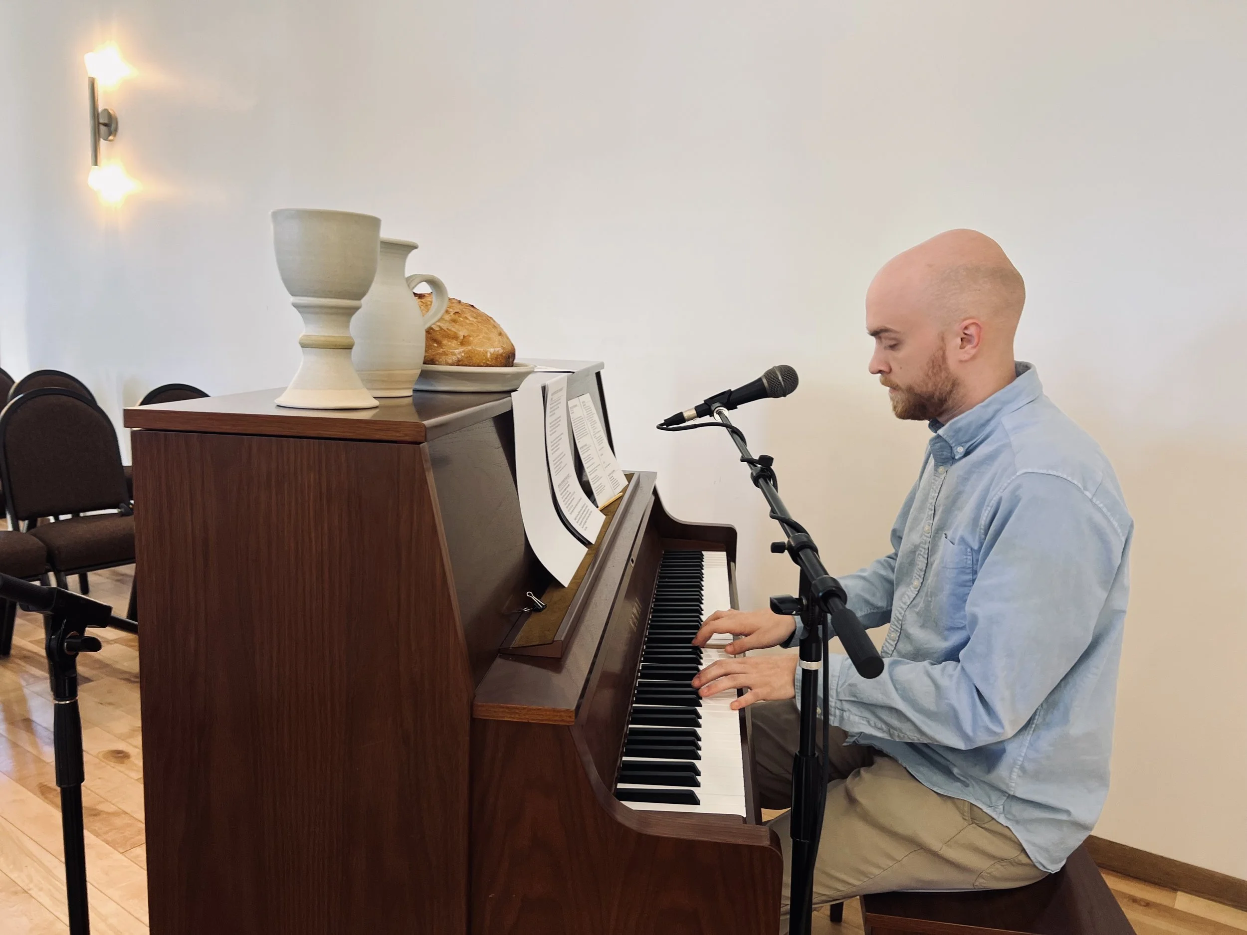 A man with a shaved head and beard, wearing a light blue shirt, is sitting and playing a brown upright piano. There is a microphone in front of him. On top of the piano, there are large ceramic vases and a loaf of bread on a plate. The room has a plain white wall and several brown chairs.