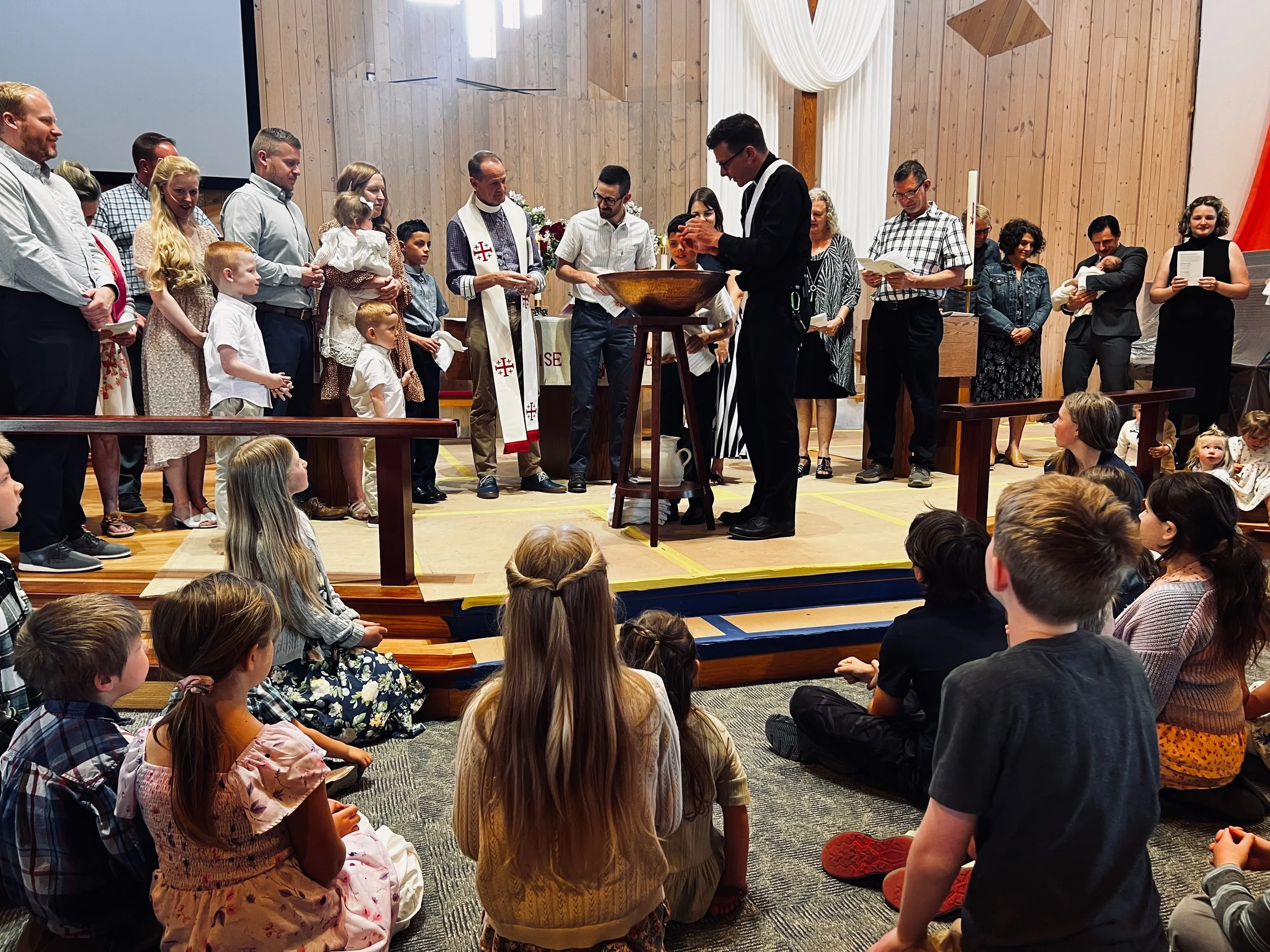 A group of people gathered in a church for a baptism ceremony. Children are seated on the floor, and adults are standing, watching the baptism.