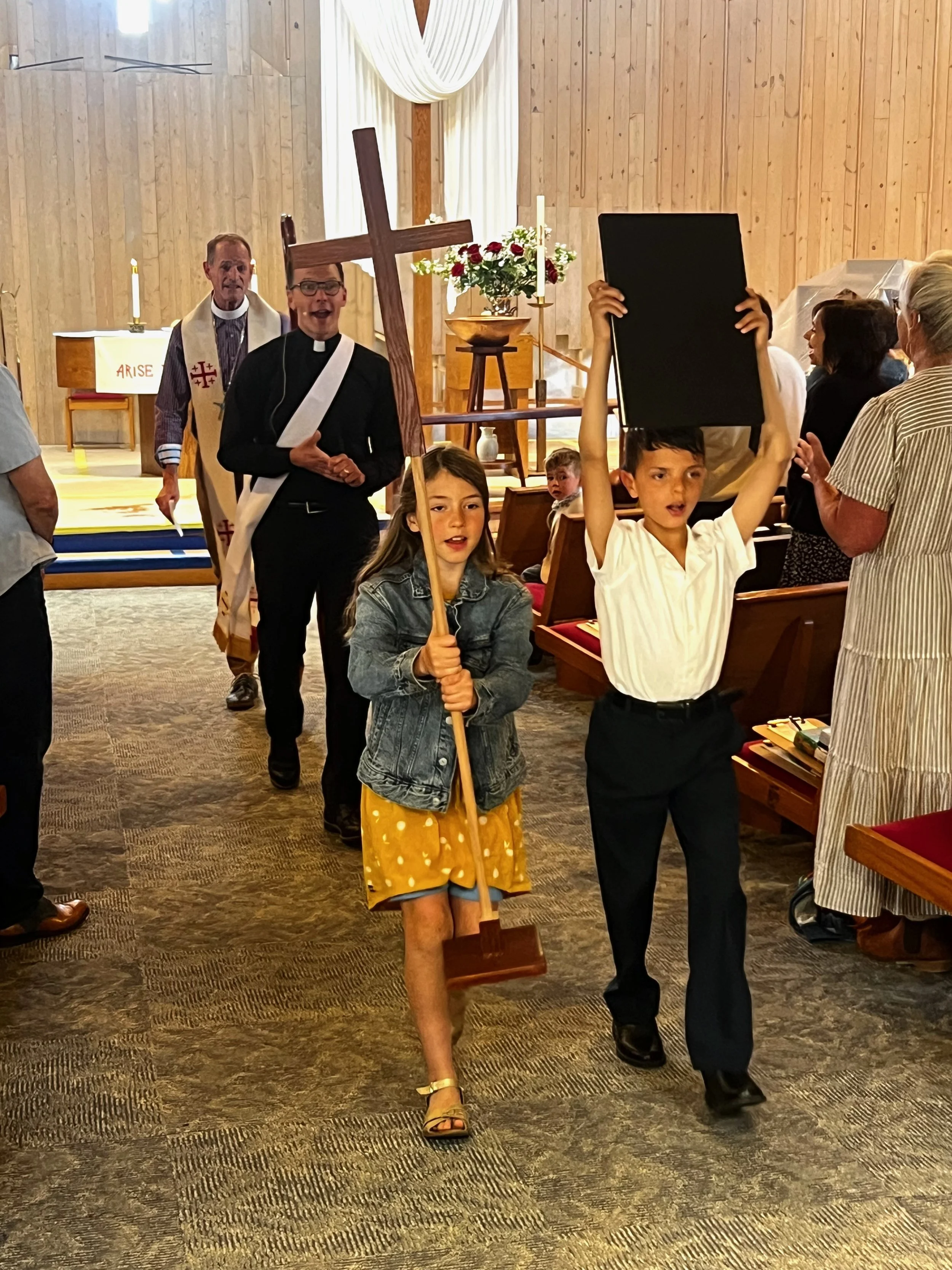 A church service with children leading a procession, including a girl in a yellow skirt holding a cross and a boy holding a framed picture, while the congregation watches.
