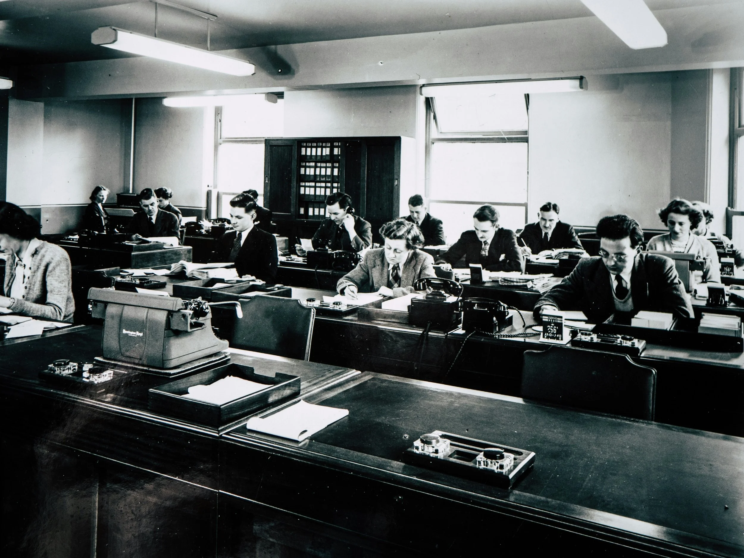 Black and white photo of a mid-20th century office workspace with women and men working at desks with typewriters and papers.