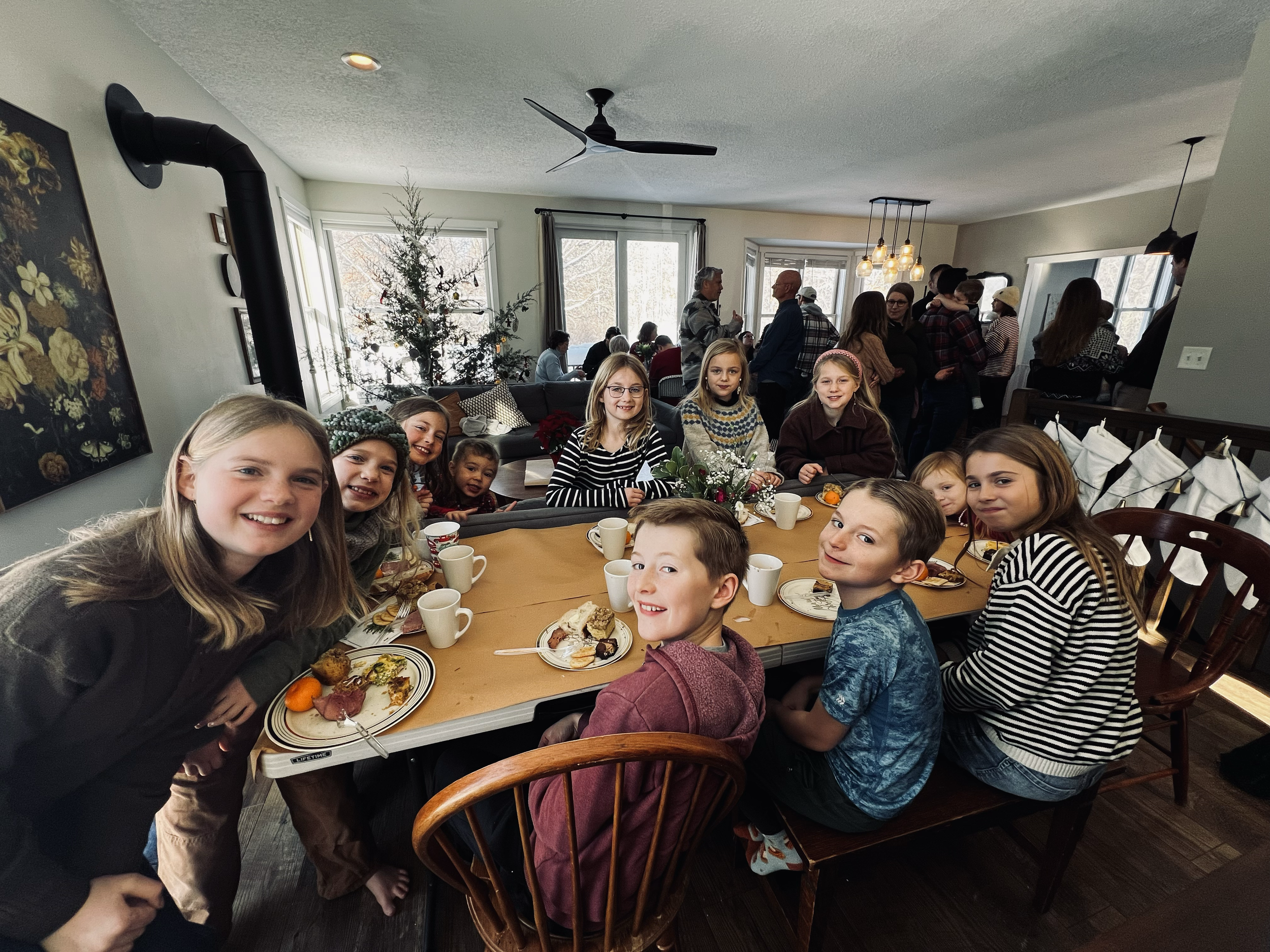 Children and adults gathered around a table with food, celebrating a holiday inside a home decorated with a Christmas tree.