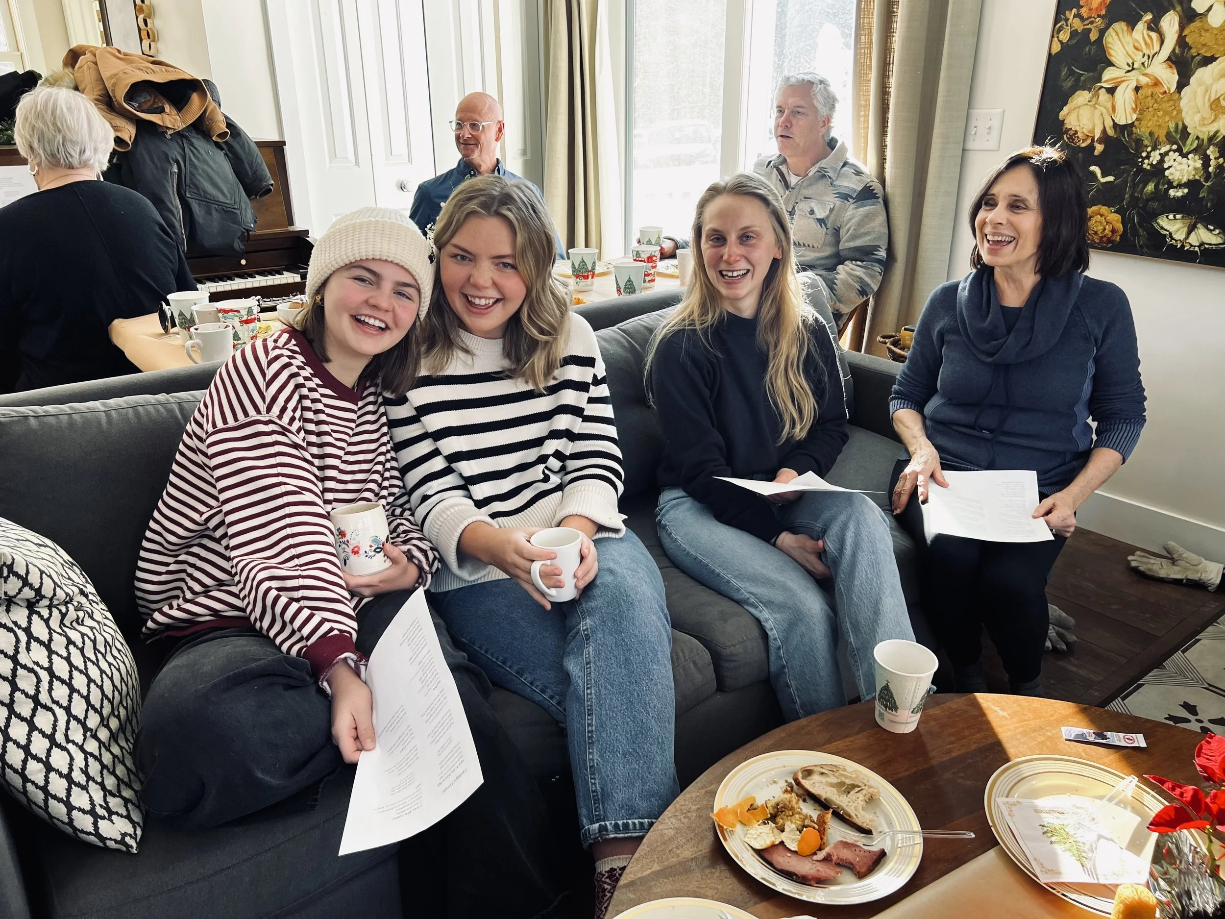 Group of four women laughing and sitting on a couch at a holiday gathering, with a table of food in front of them and other people in the background.