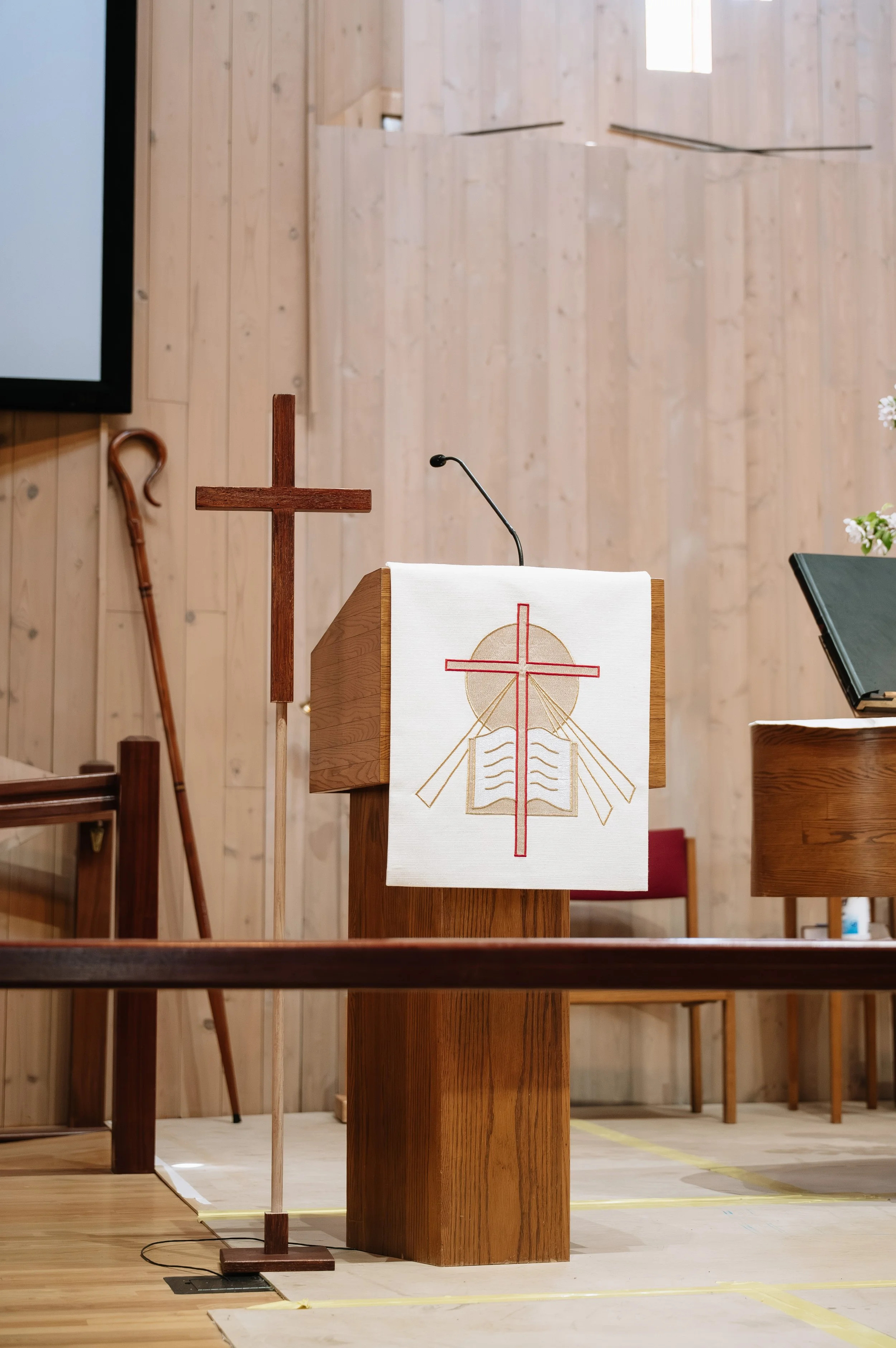 Inside a church, wooden altar with a white cloth featuring a red cross and an open book with a gold circle, a wooden cross on a stand, a lectern, and a music stand. The background has light wooden paneling and a small window.