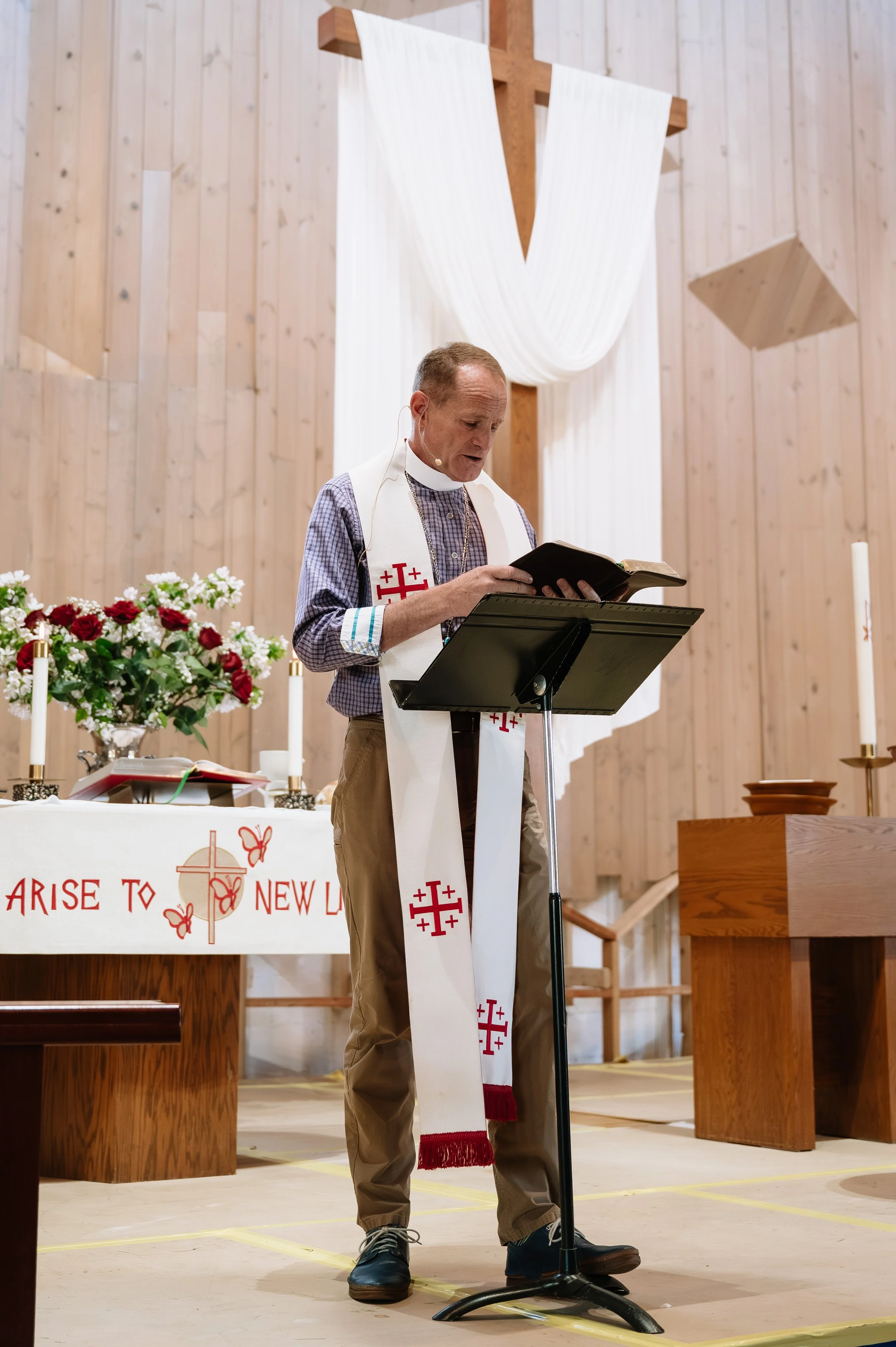 A man standing at a lectern inside a church or chapel, reading from a book, with an altar decorated with flowers and candles behind him and a large wooden cross with white drapery hanging in the background.