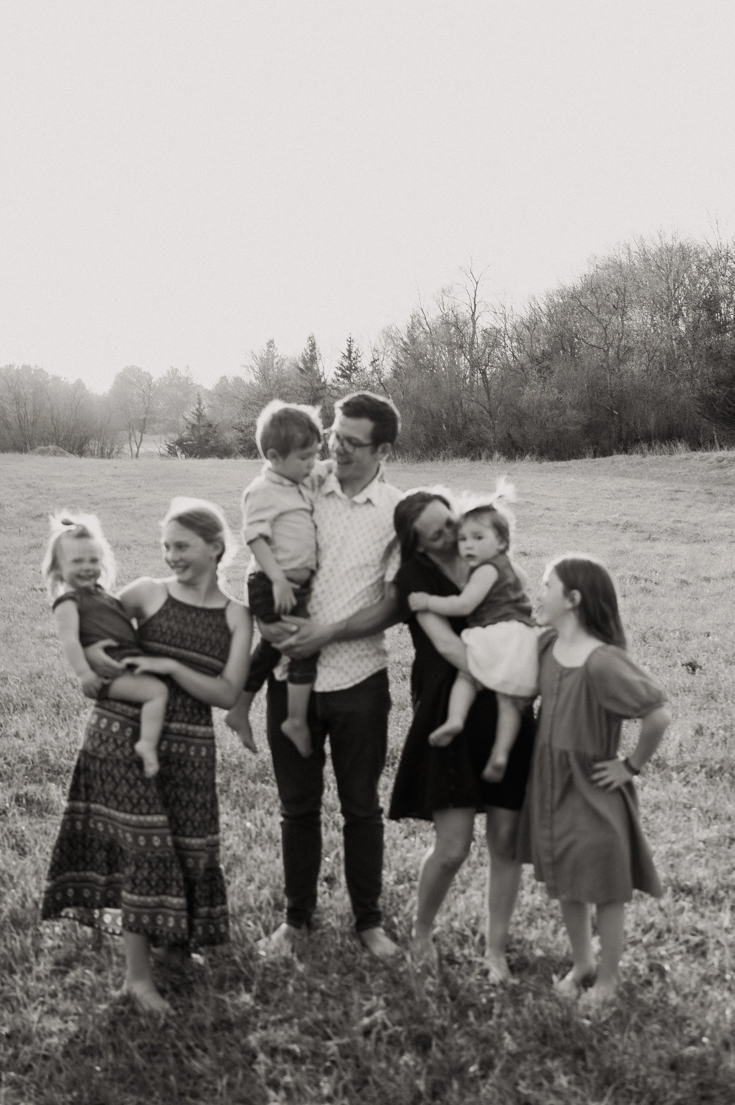 A black-and-white photo of a group of children and adults outdoors in a grassy field, smiling and interacting, with trees and open sky in the background.