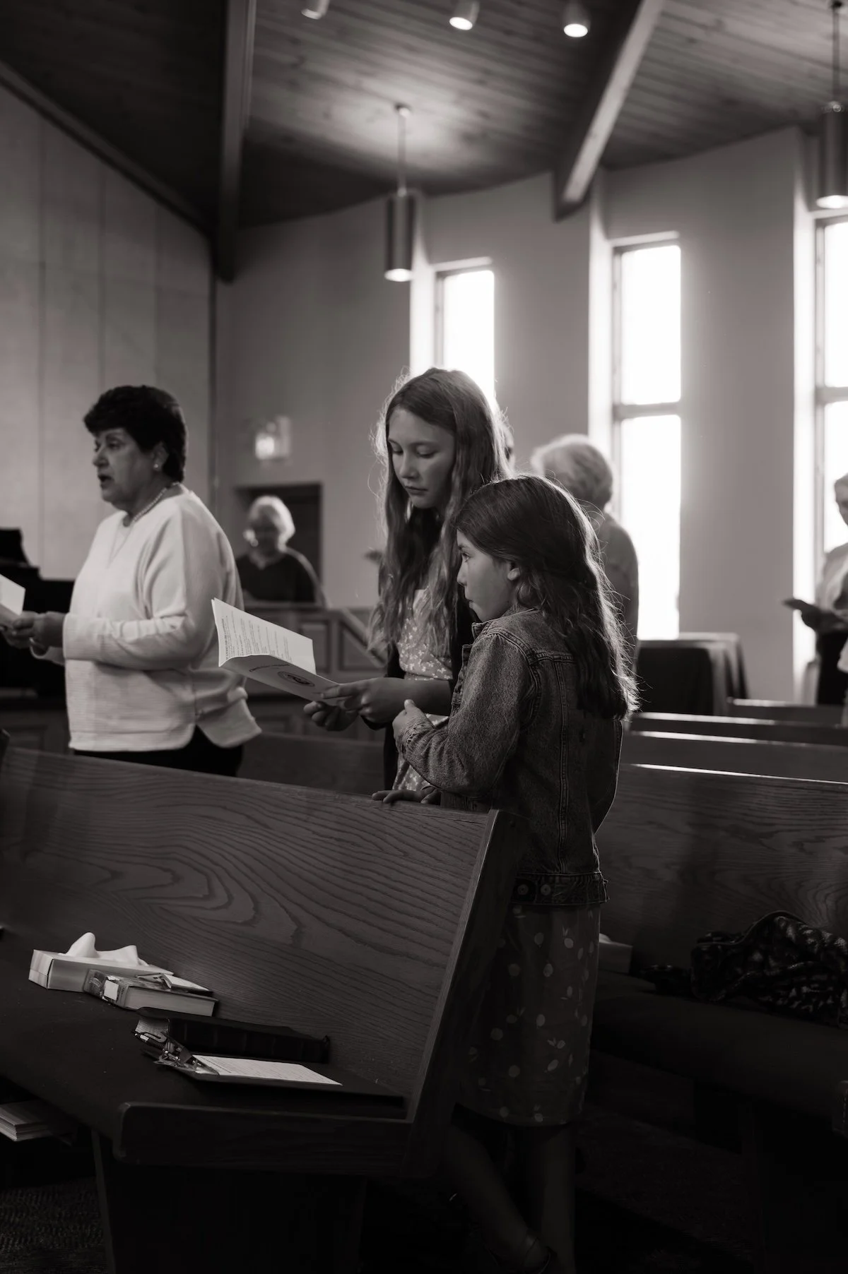 People standing and reading in a church or meeting hall, with sunlight coming through windows in the background, including an older woman and a young girl in the foreground.