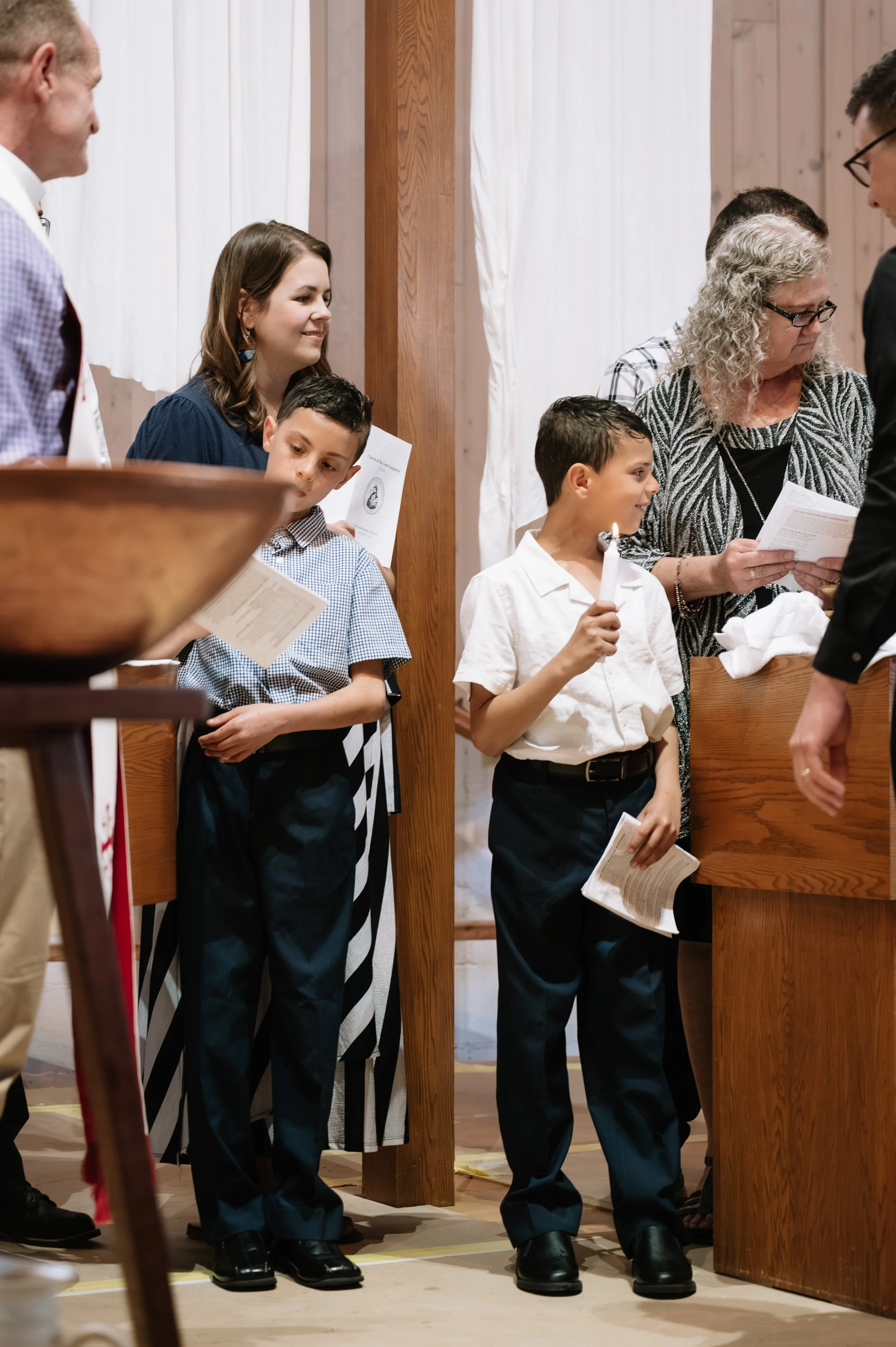 Children and adults participating in a religious or ceremonial gathering inside a church or chapel, some holding prayer books and one holding a candle.