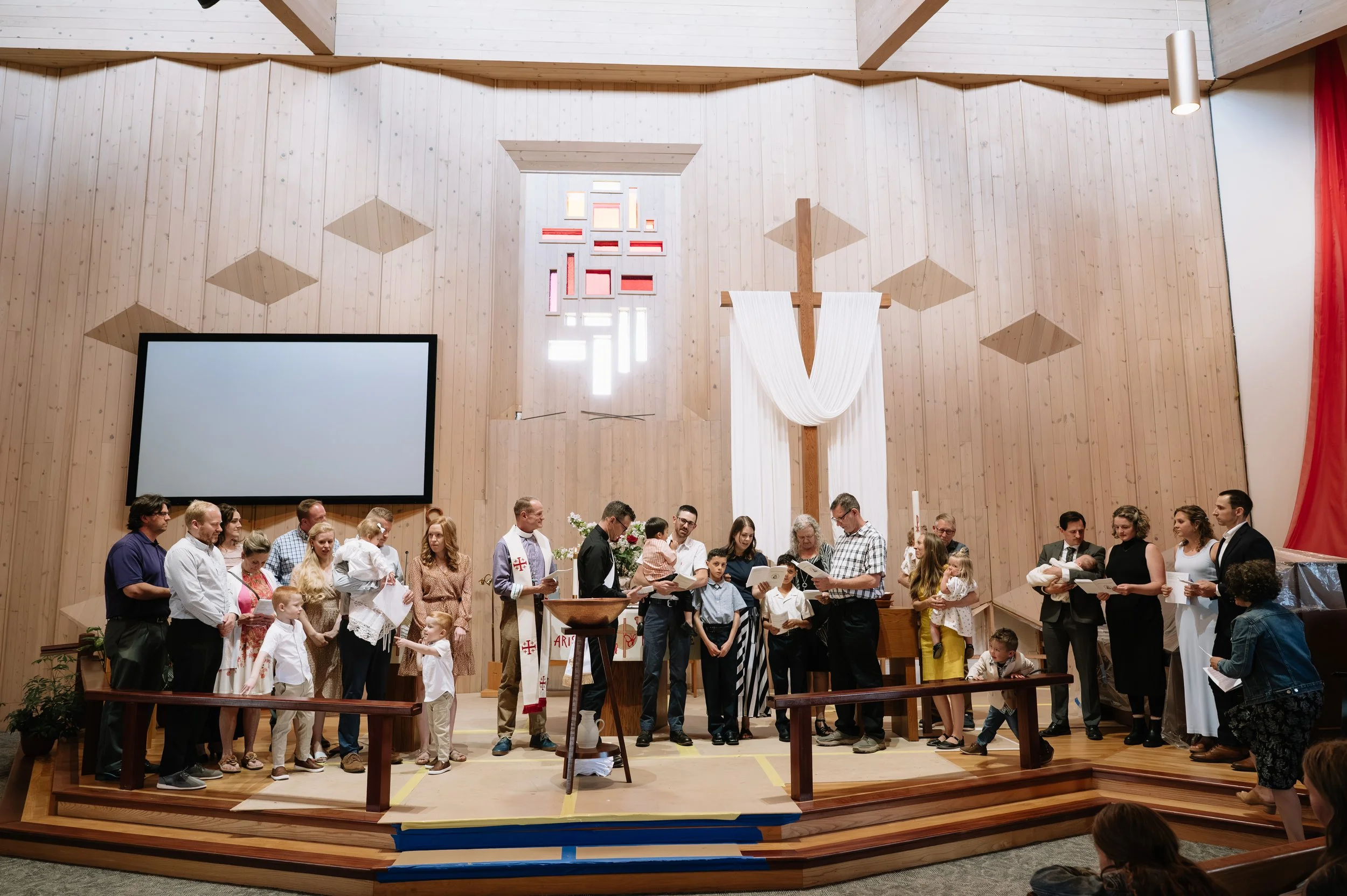 People gathered inside a church for a religious service or ceremony, with a wooden altar, cross, and stained glass window in the background.