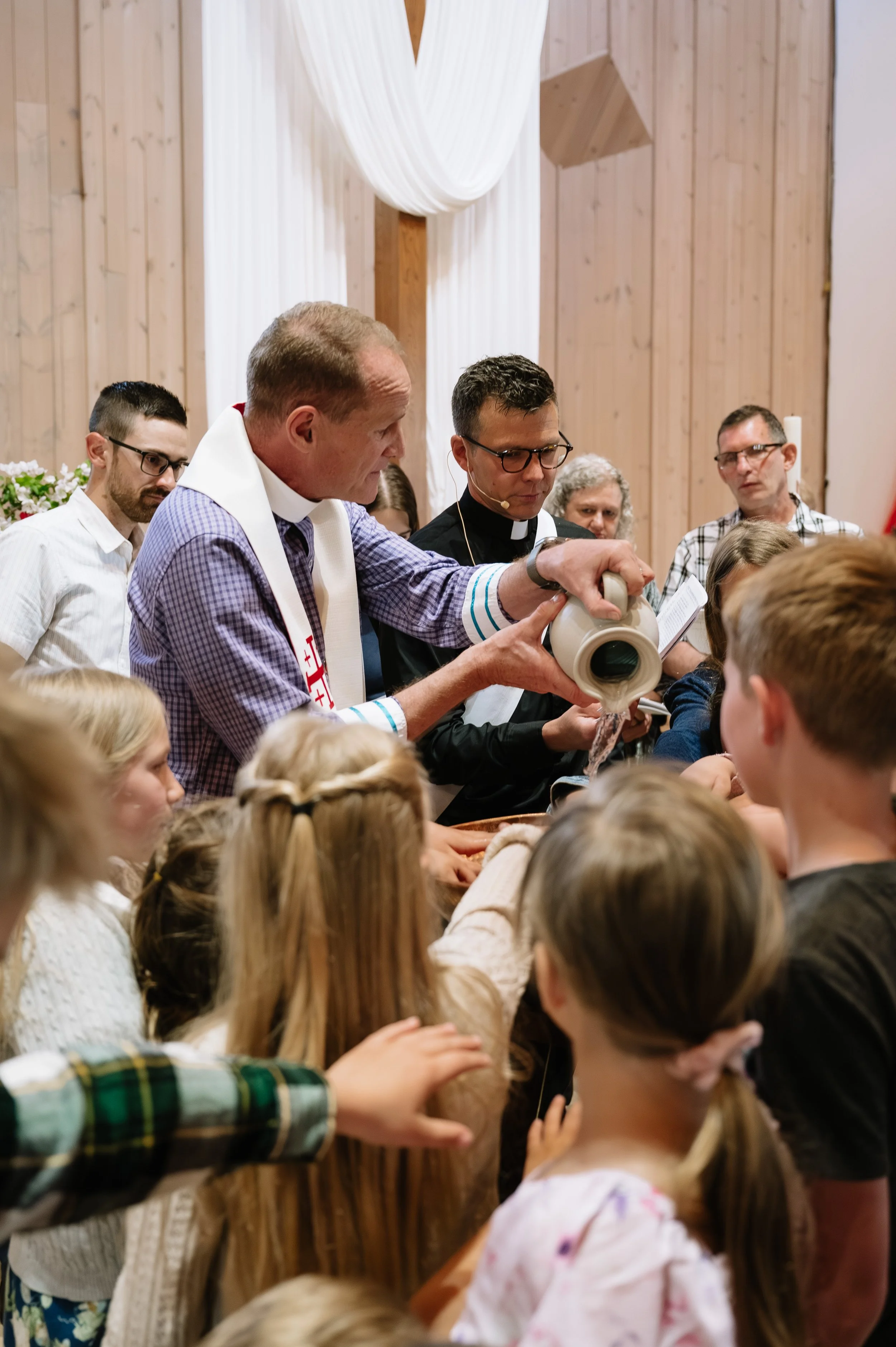 A religious ceremony with a priest and a minister performing a ritual involving pouring water on people's heads, with children and adults gathered closely around.